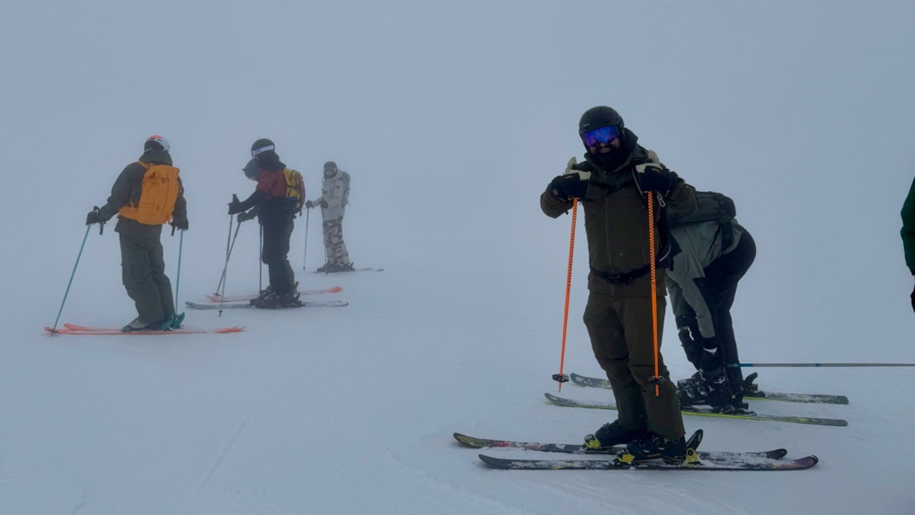 A group of skiers of varying abilities pause together on a snowy slope in mist