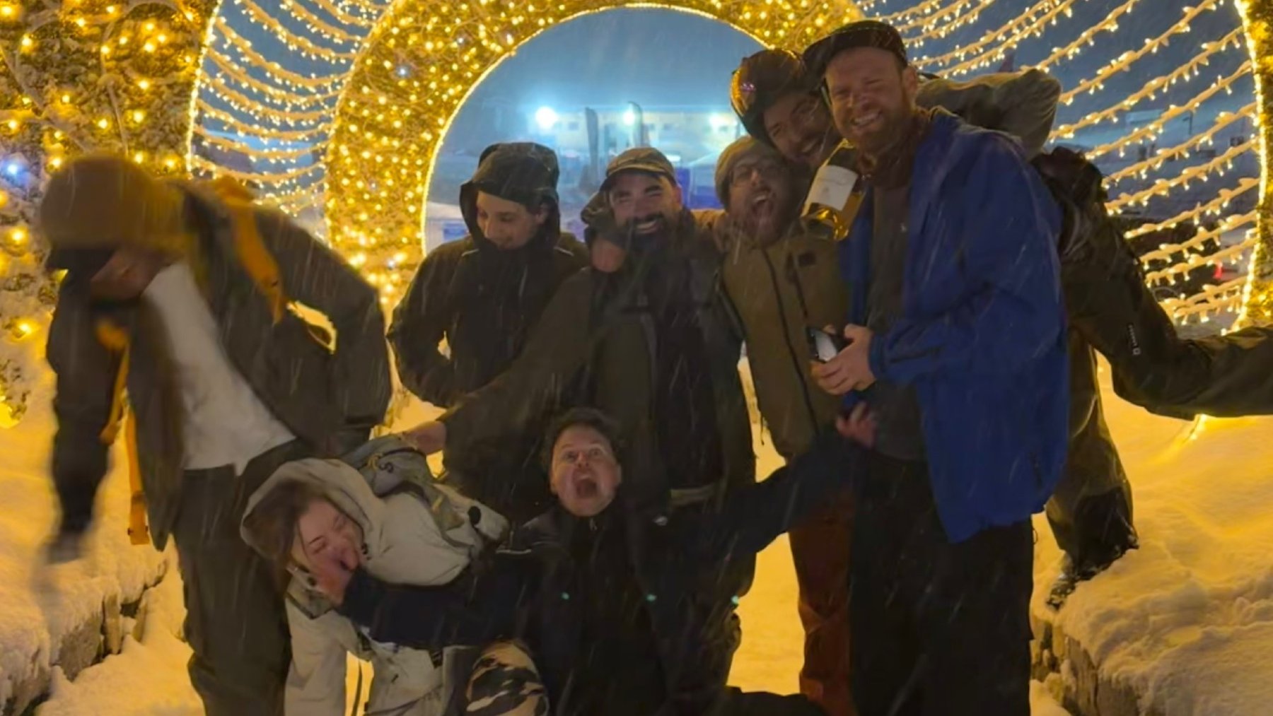 A group of friends in ski gear pose in the snow beneath a brightly lit archway.