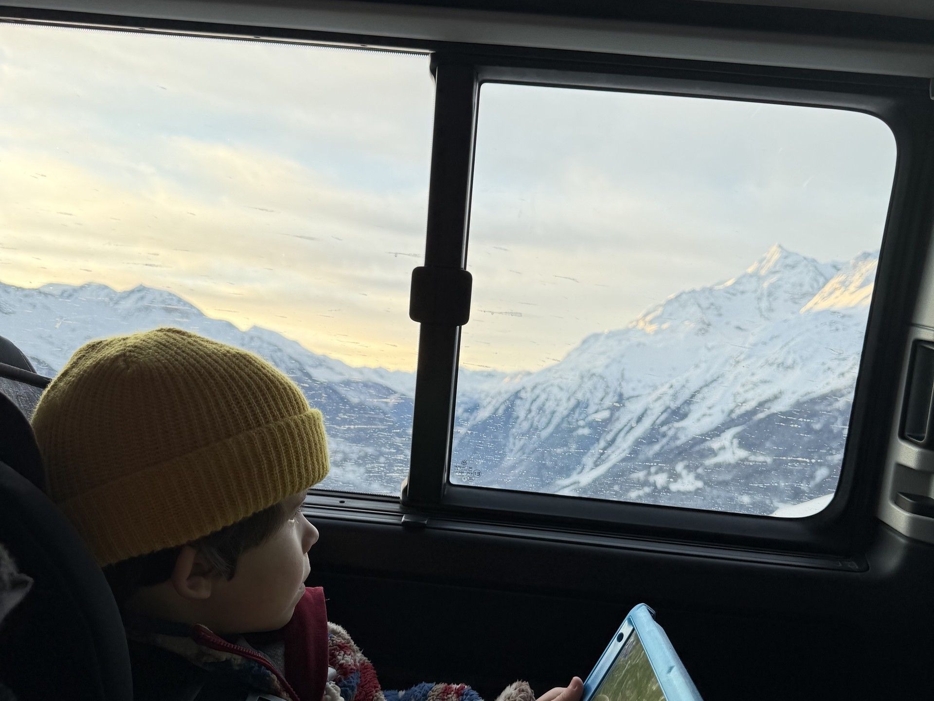 Child in a ski trip car seat playing with a tablet while mountains snow peaks outside window