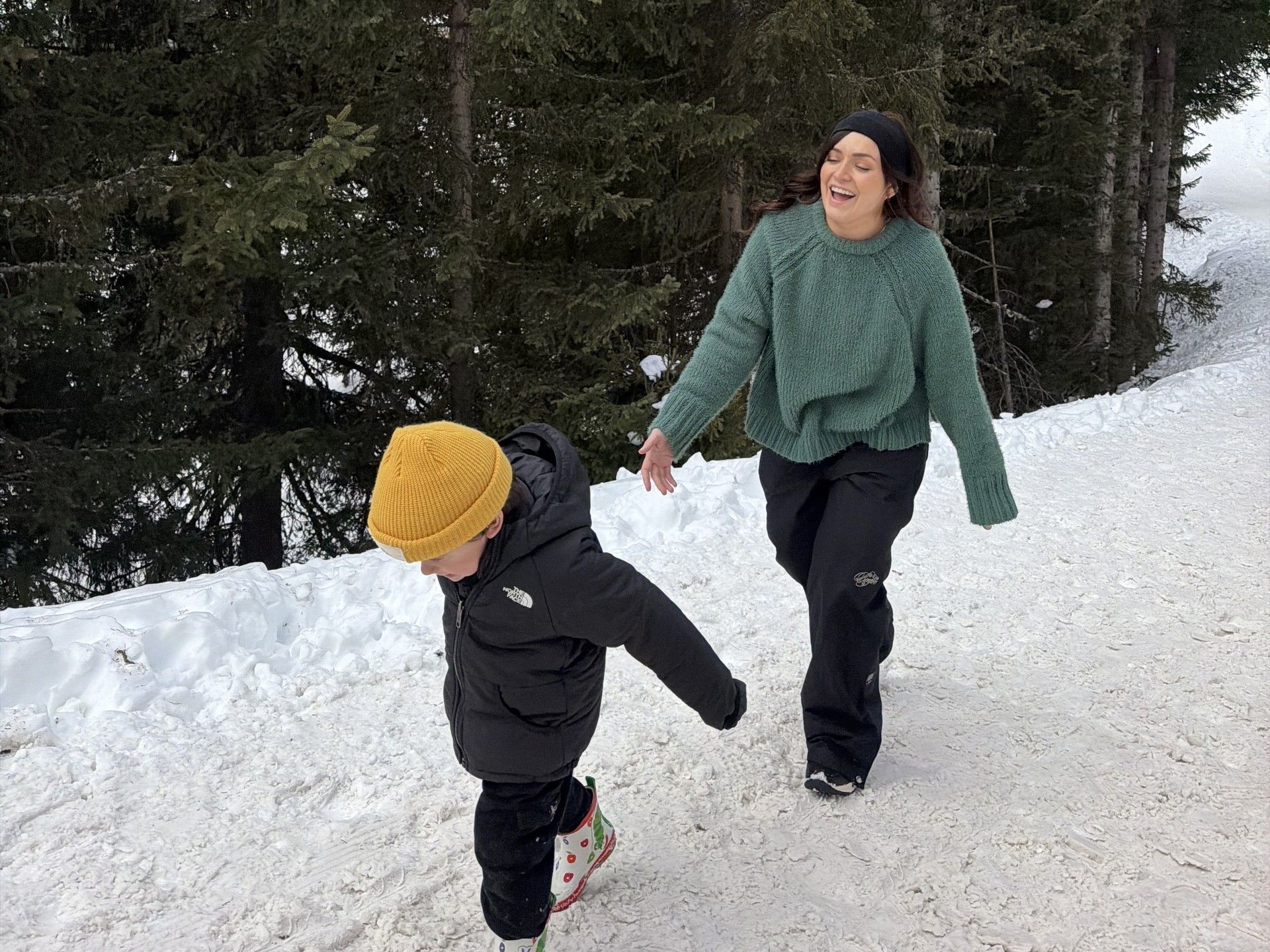 Two people walking downhill in snow on skis in a forested ski area, with one child leading