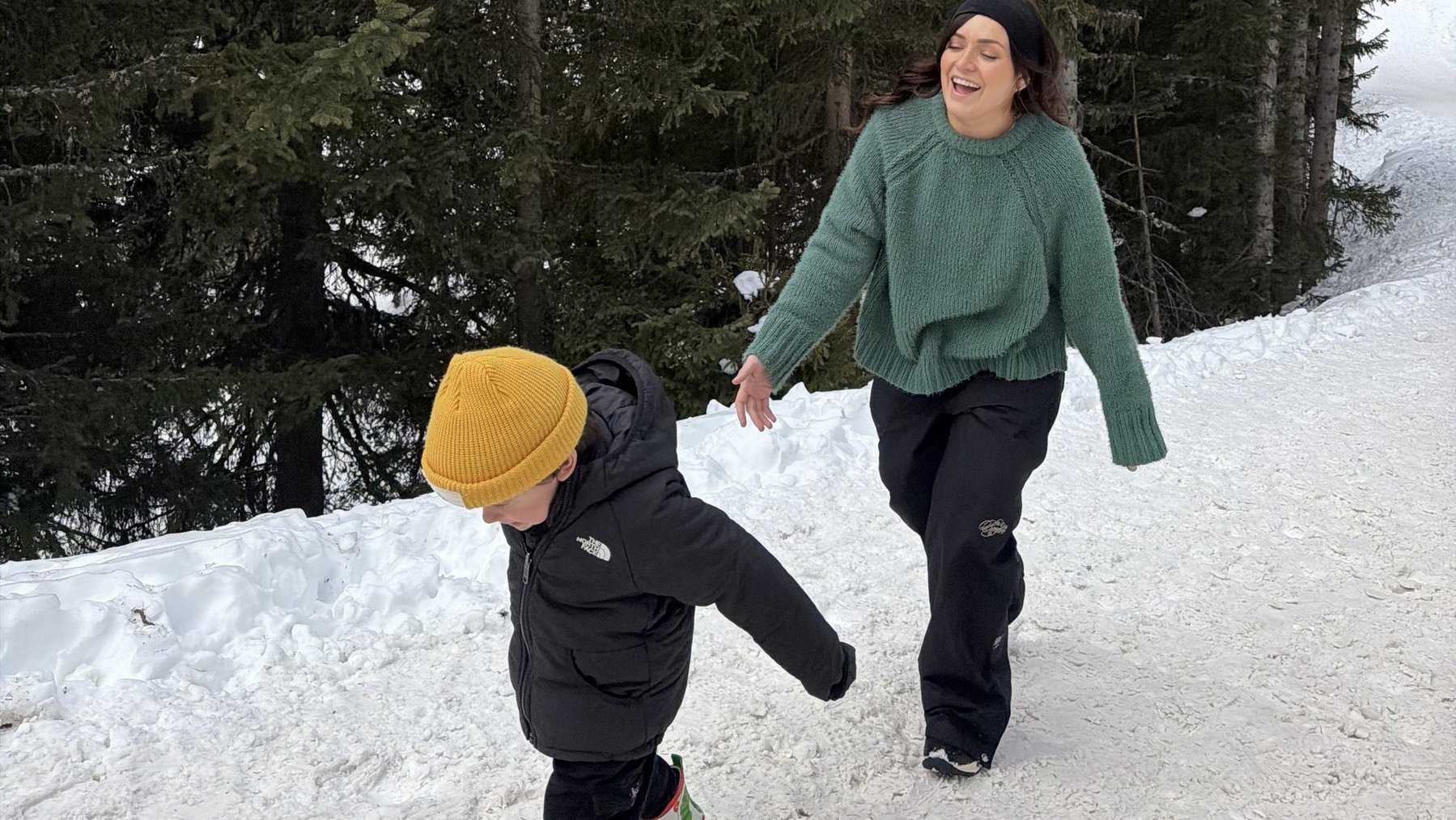 Two people walking downhill in snow on skis in a forested ski area, with one child leading