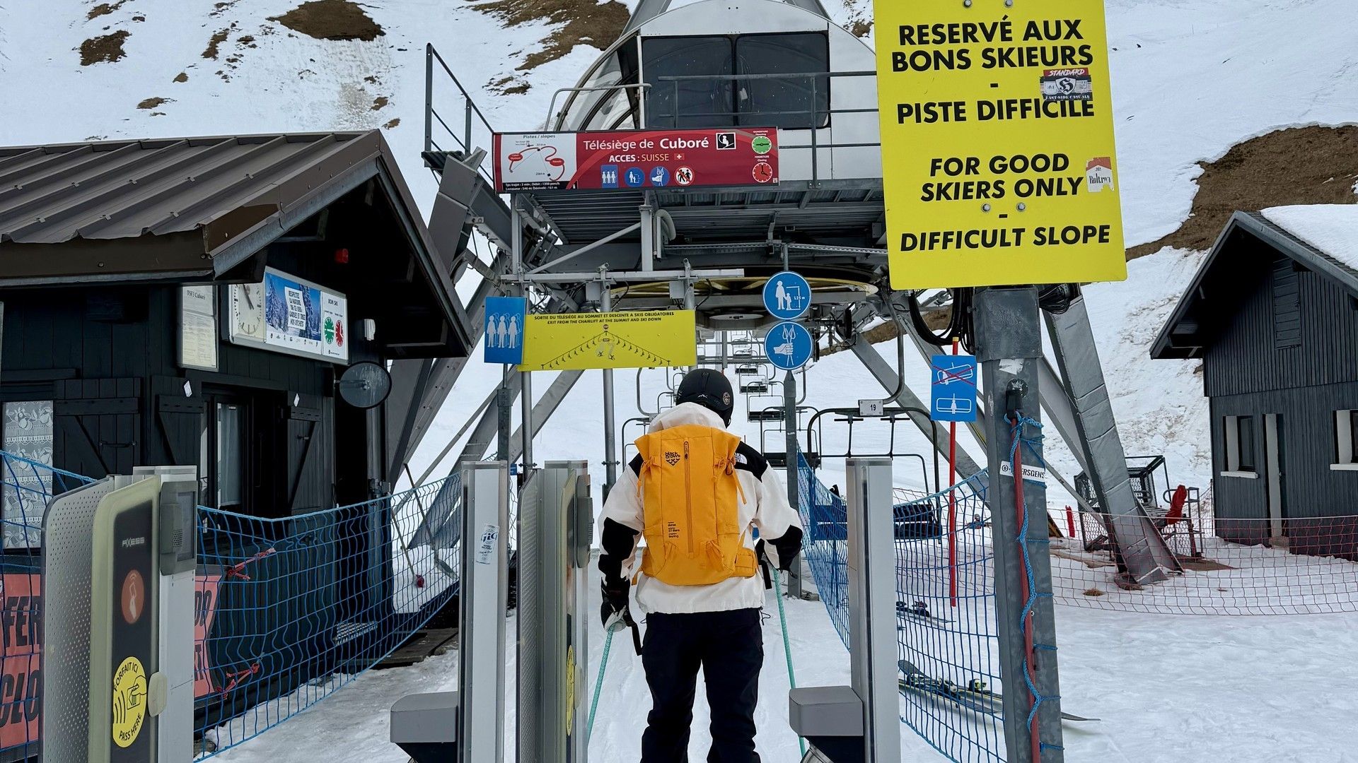 Ski lift access gate with warning signs in snowy resort, skier walking uphill toward the lifts