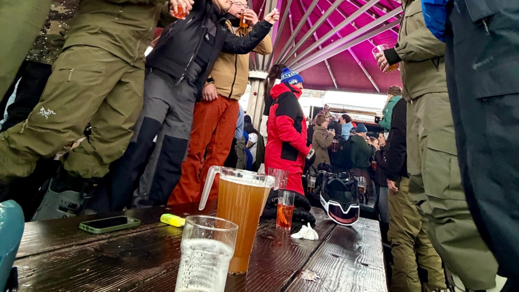 Skiers in winter gear gather around a table with beer glasses at après-ski beneath a red canopy.