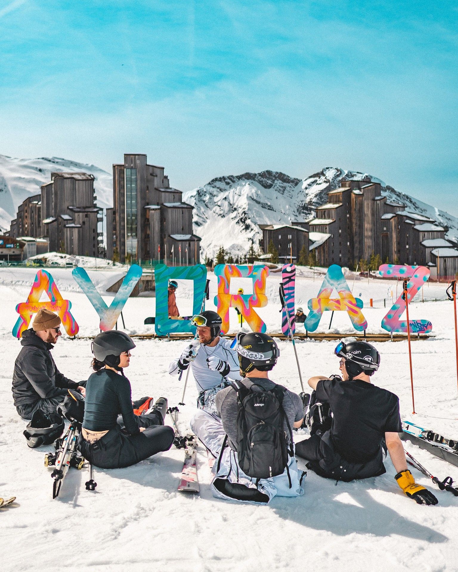 Ski instructors and skiers in helmets gather on snow with colorful pole covers near alpine resort buildings