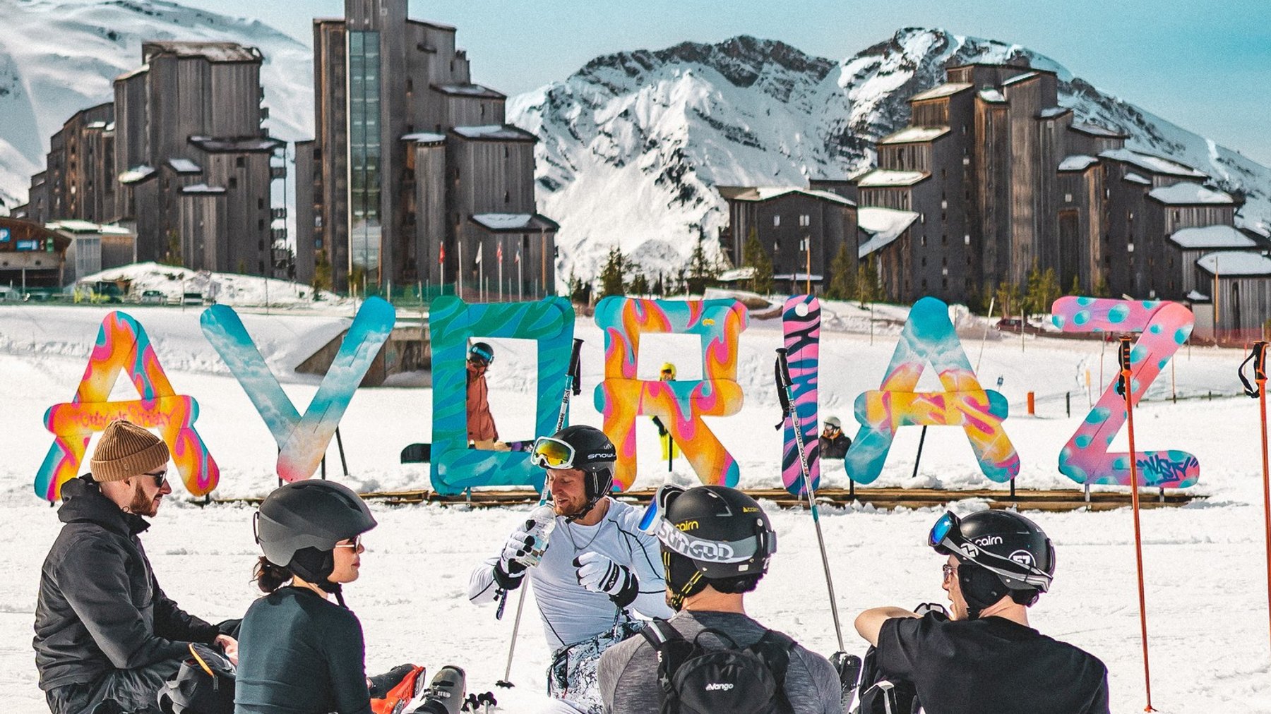 Ski instructors and skiers in helmets gather on snow with colorful pole covers near alpine resort buildings