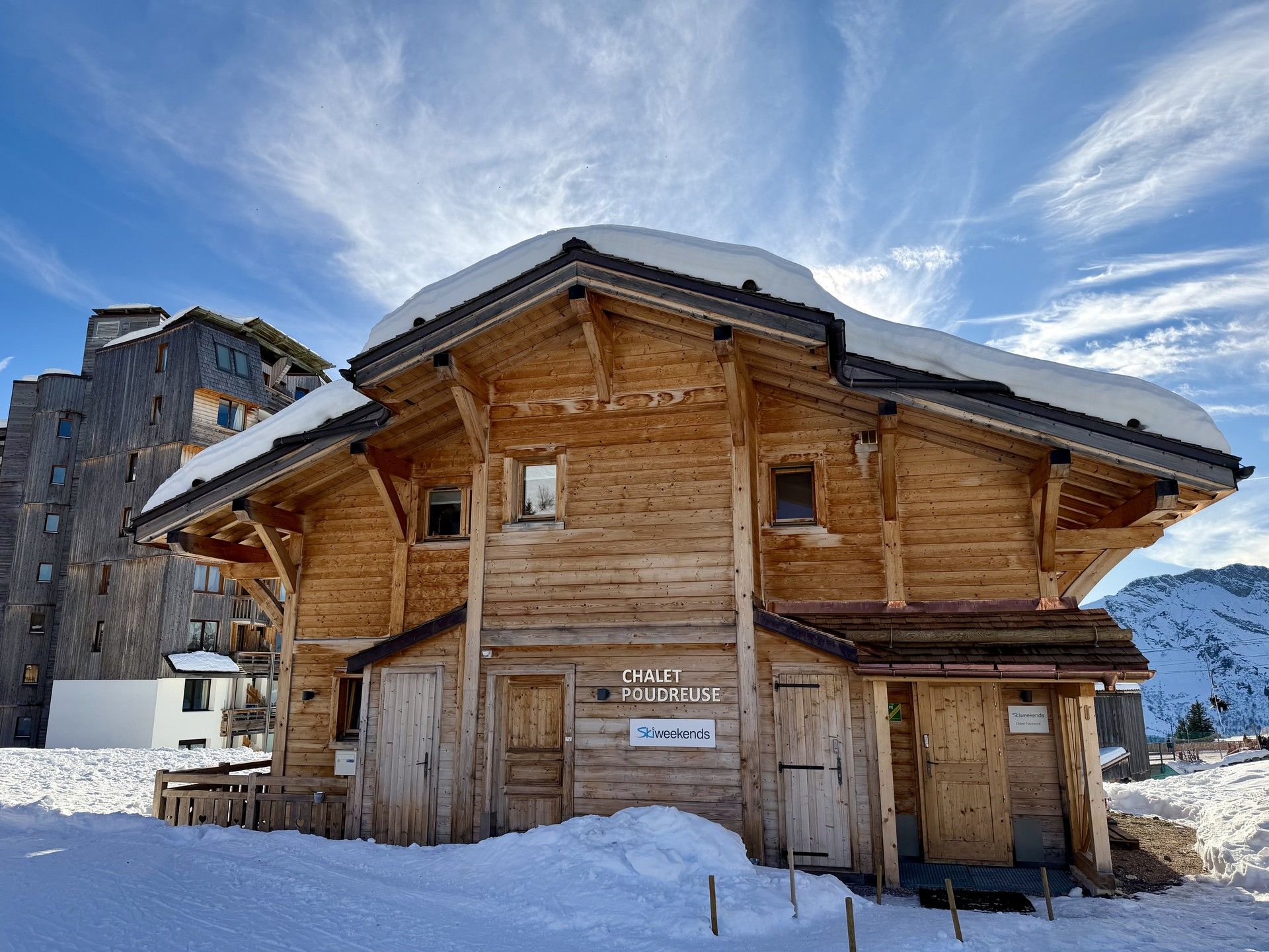 Wooden ski chalet covered in snow with alpine mountains and blue sky in the background
