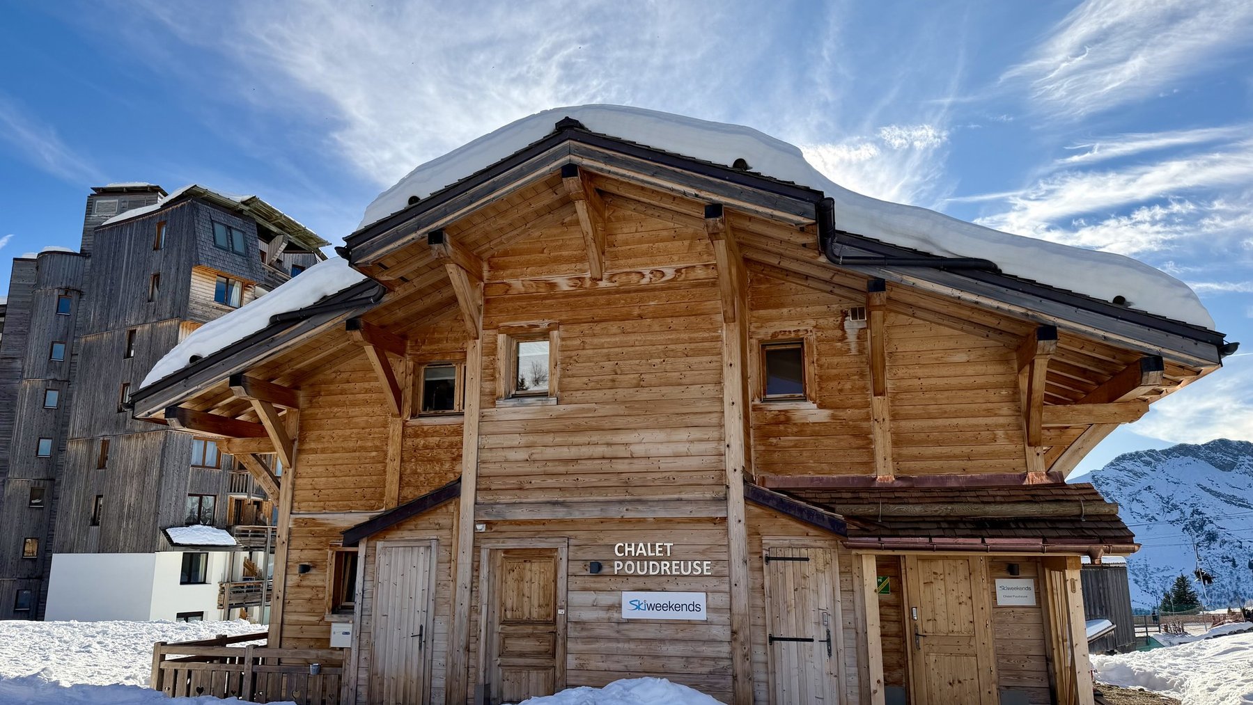 Wooden ski chalet covered in snow with alpine mountains and blue sky in the background