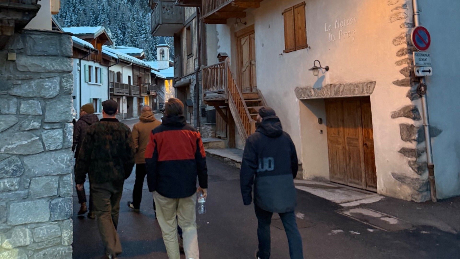 Three friends walk through a snowy Alpine village decorated with festive lights and backed by a forested mountain.