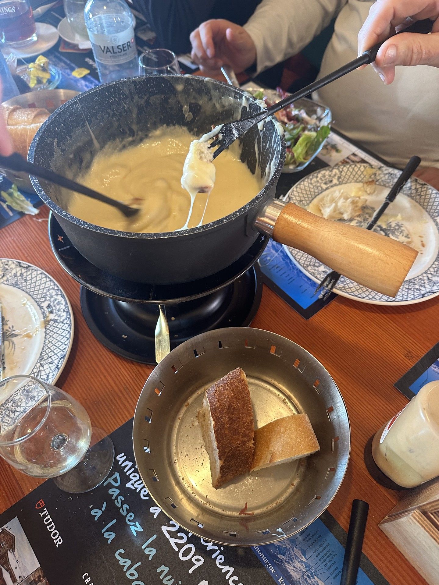 Chef stirring a cheese fondue pot at a ski resort dining table, with bread and plates nearby