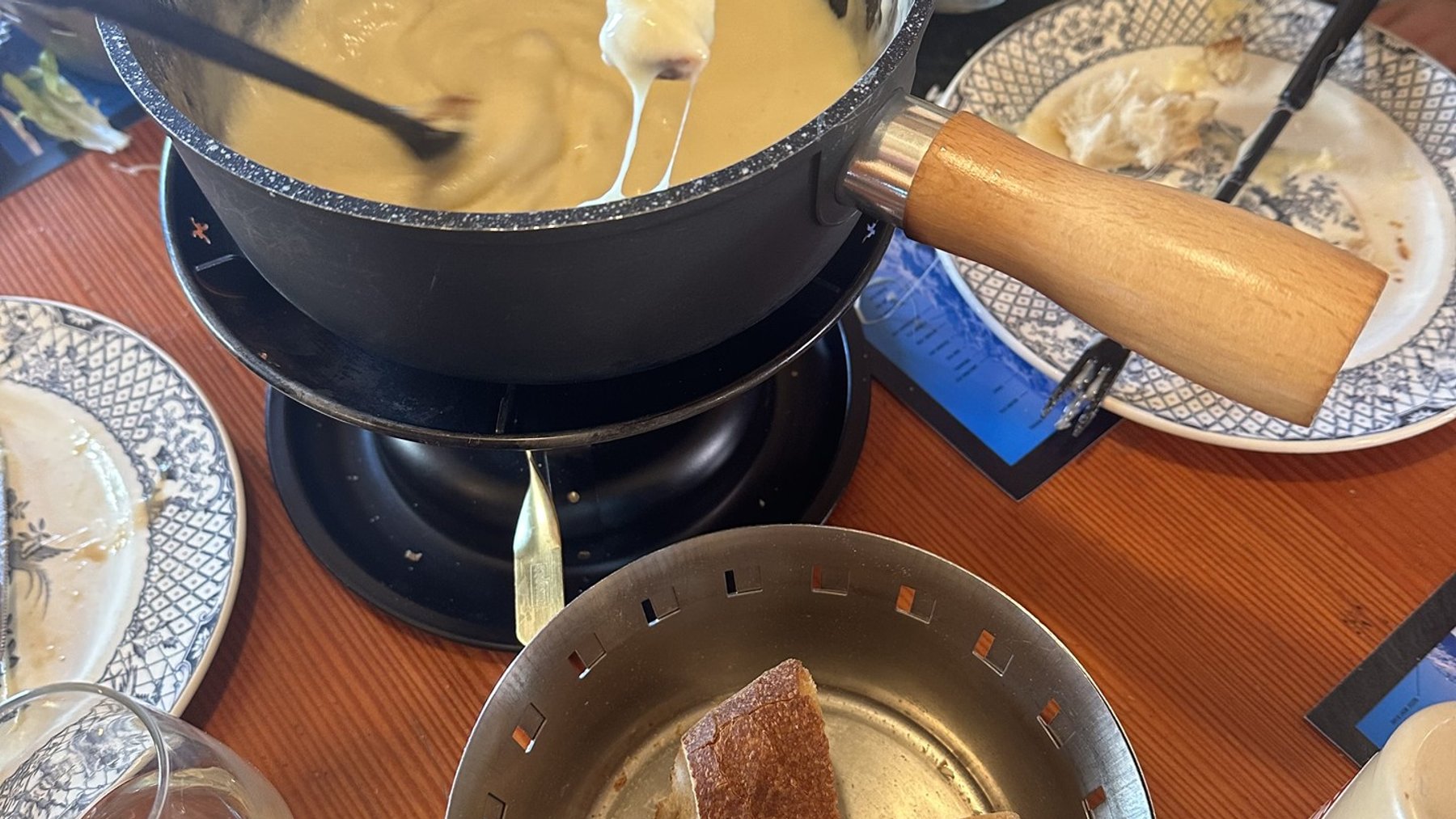 Chef stirring a cheese fondue pot at a ski resort dining table, with bread and plates nearby