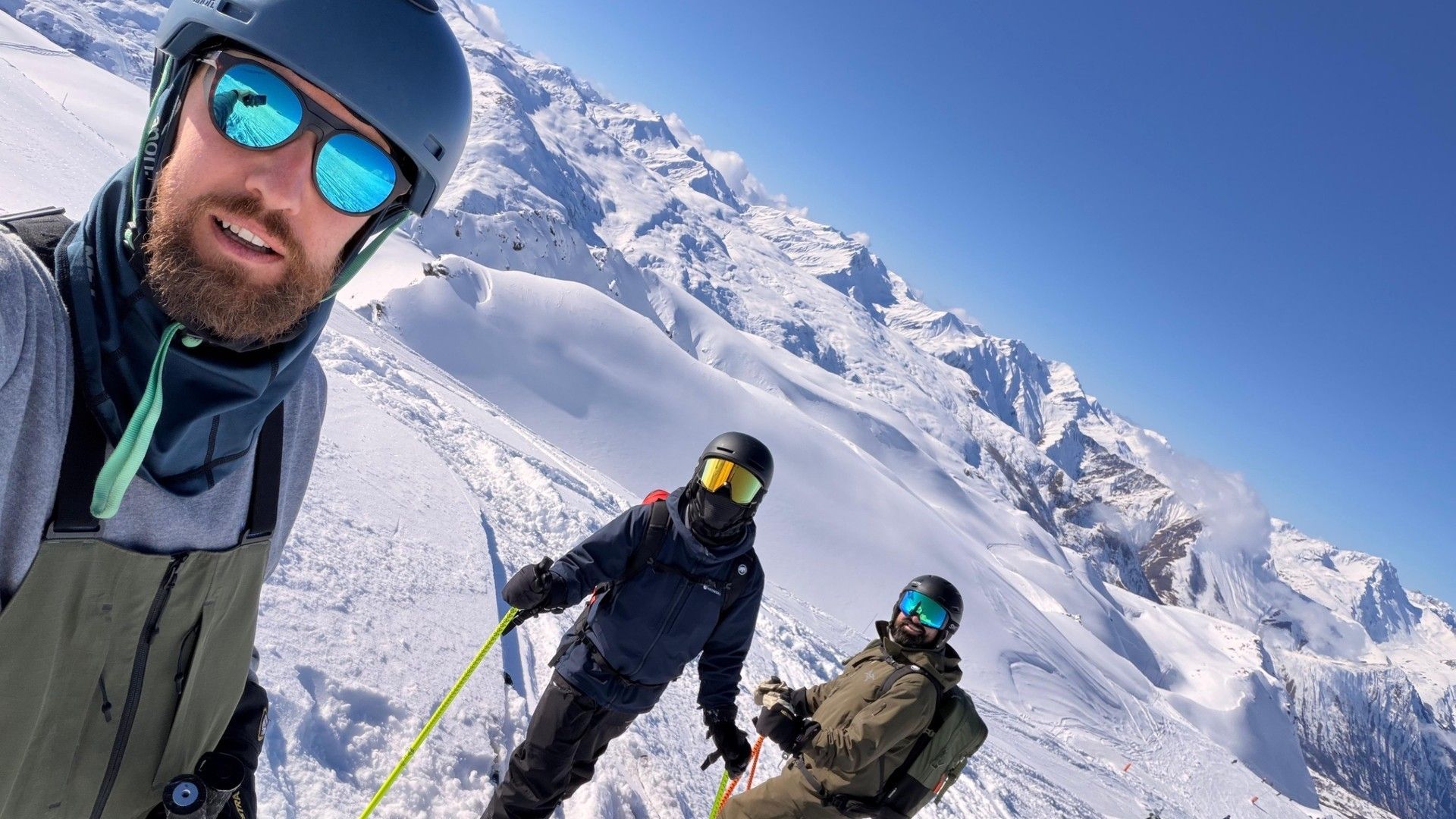 Three skiers pose on a snowy alpine slope, with a mountain range behind them