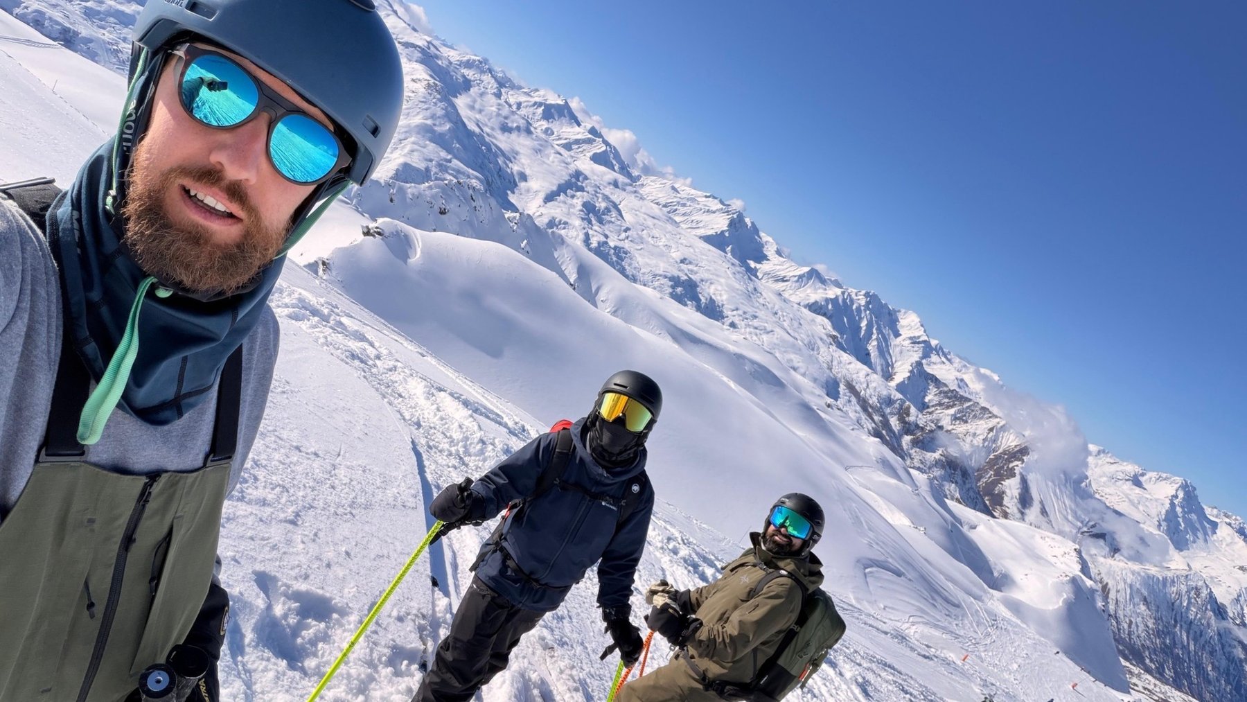 Three skiers pose on a snowy alpine slope, with a mountain range behind them