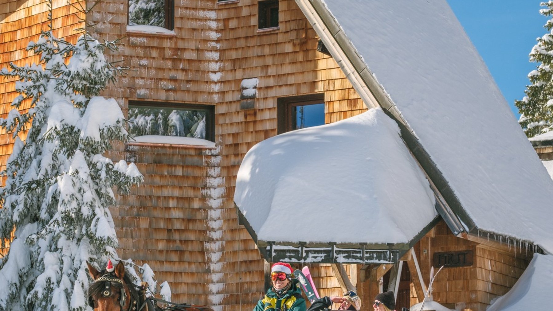 Horse-drawn sleigh rides past a snow-covered wooden lodge on a sunny winter day