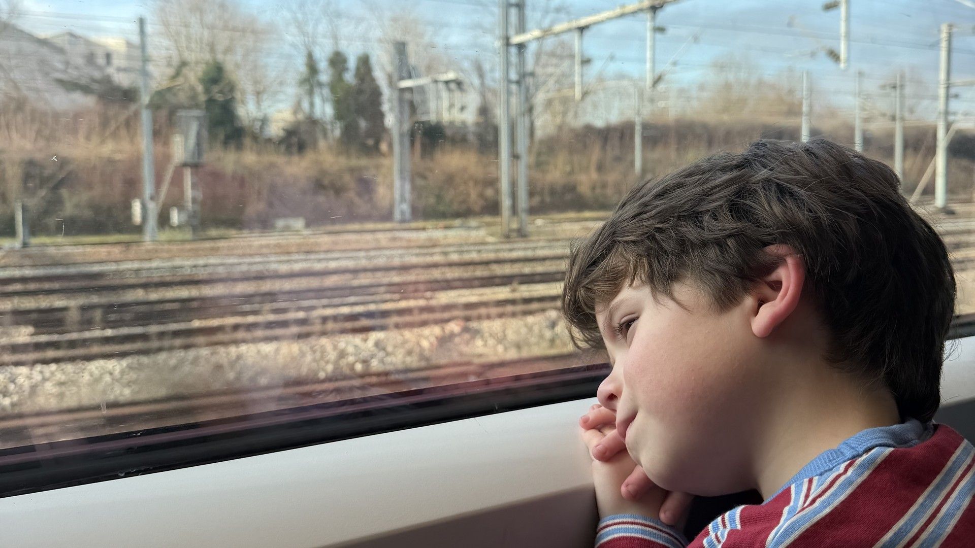 Child gazes out a train window at tracks and power lines during a journey.