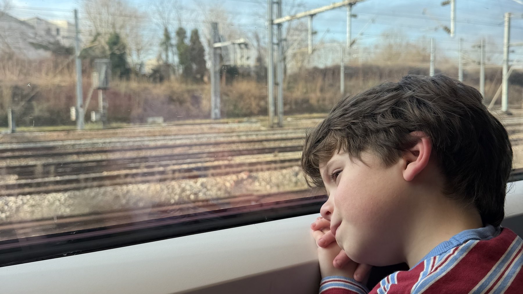 Child gazes out a train window at tracks and power lines during a journey.