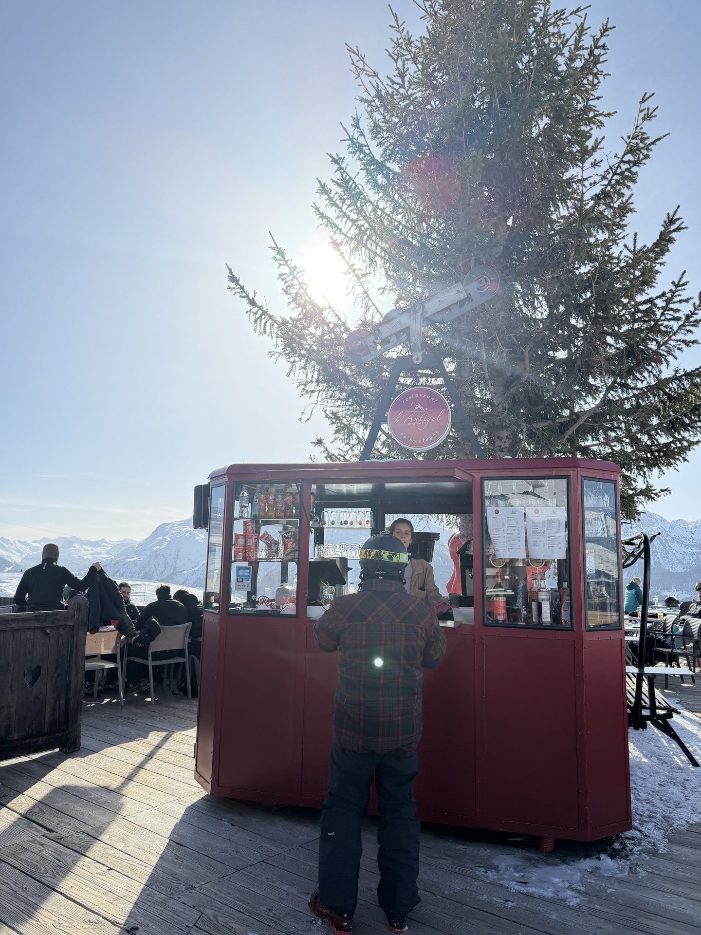 Ski lift ticket kiosk on a mountain terrace beside snow-covered benches and a tall evergreen tree