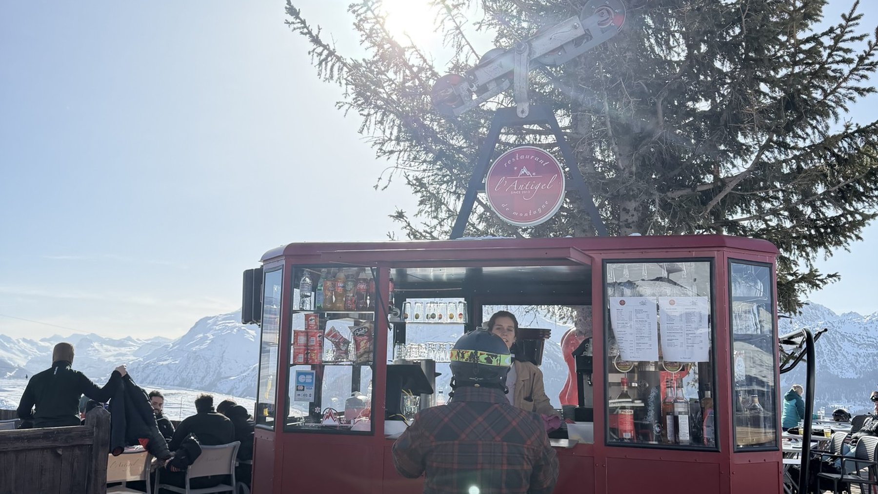 Ski lift ticket kiosk on a mountain terrace beside snow-covered benches and a tall evergreen tree