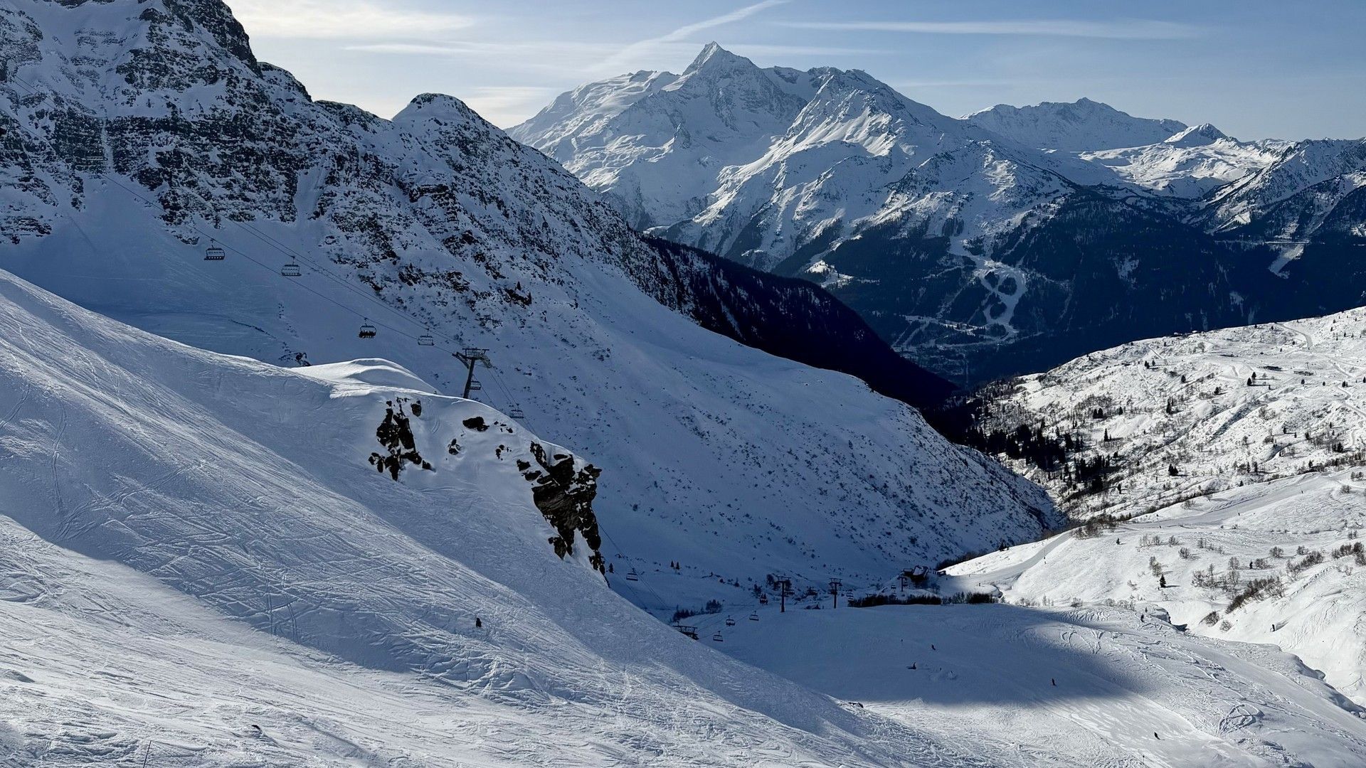 Snow-covered ski slopes and Alpine peaks under a blue sky, with a few skiers descending the foreground