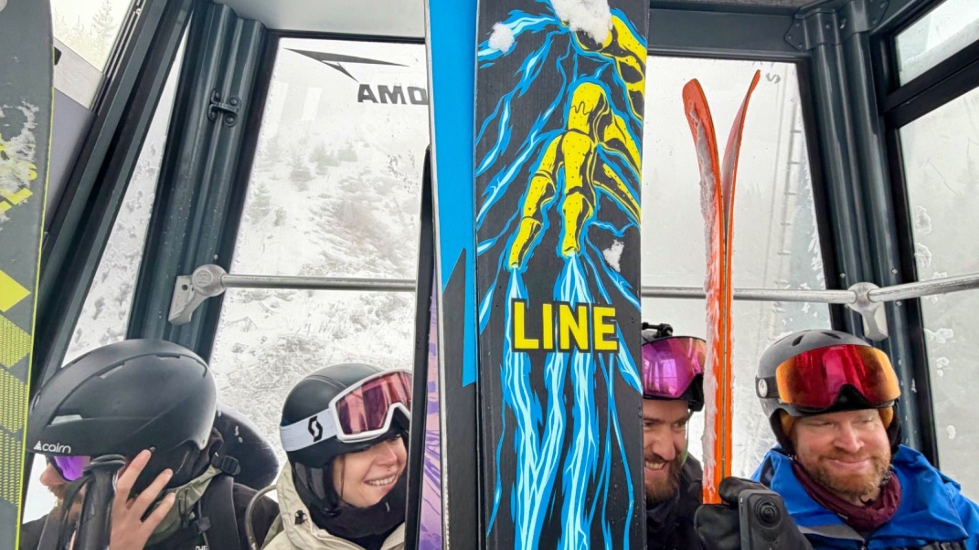 Skiers and snowboarders in helmets ride a gondola, with a LINE snowboard prominent in the foreground.