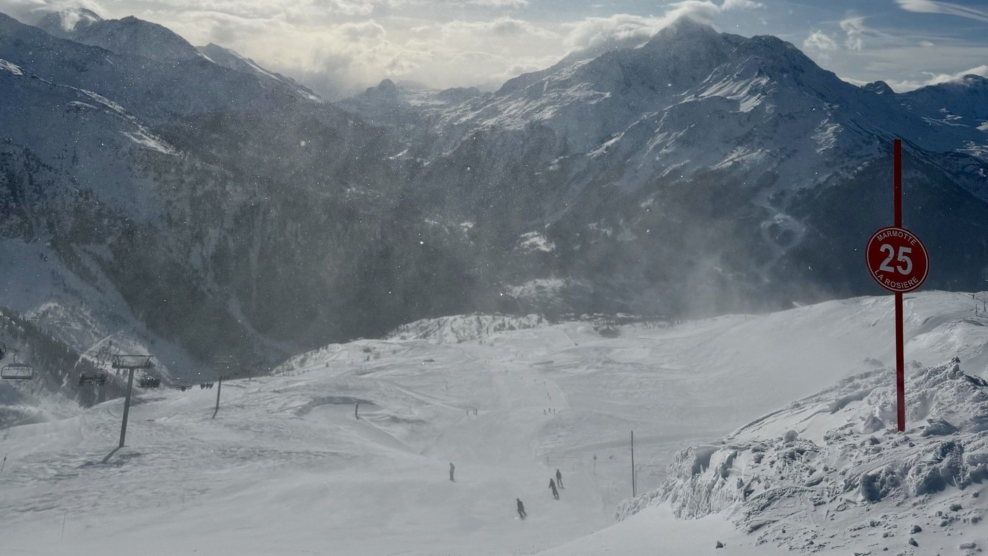 Snow-covered ski slope in La Rosière beneath the sun, with skiers descending and a red 25 sign.