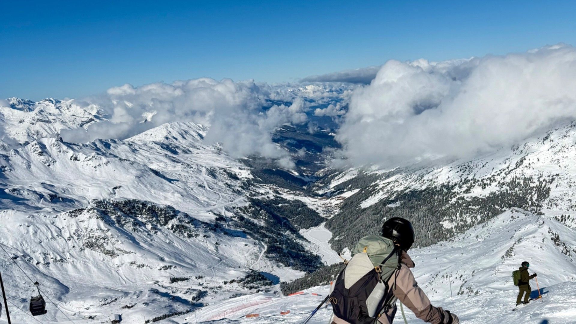 Skier descends a snowy Alpine slope above a cloud-filled valley, with another skier in the distance.