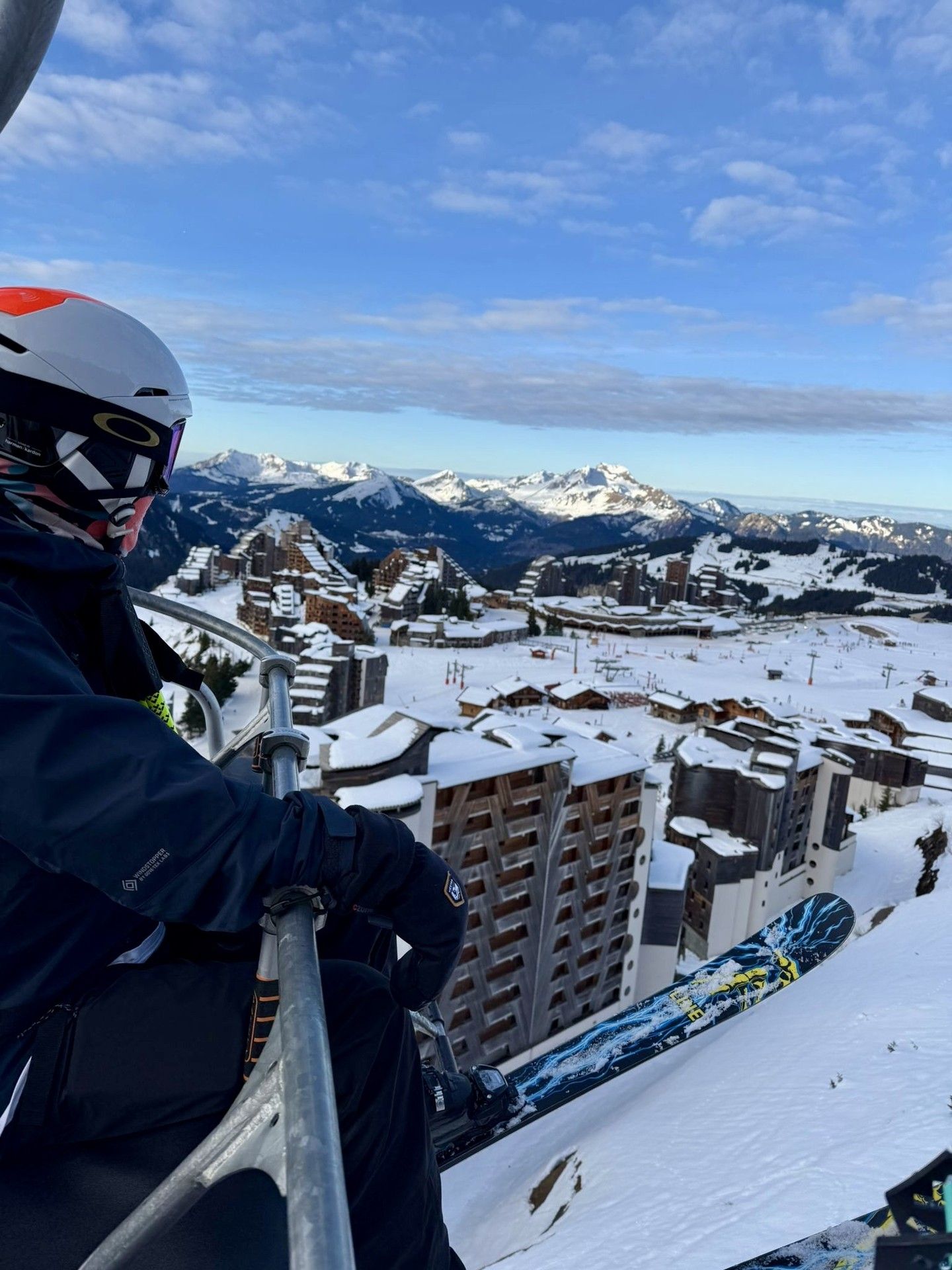 Skiing helmeted rider on chairlift with snowy mountain town and peaks in the background