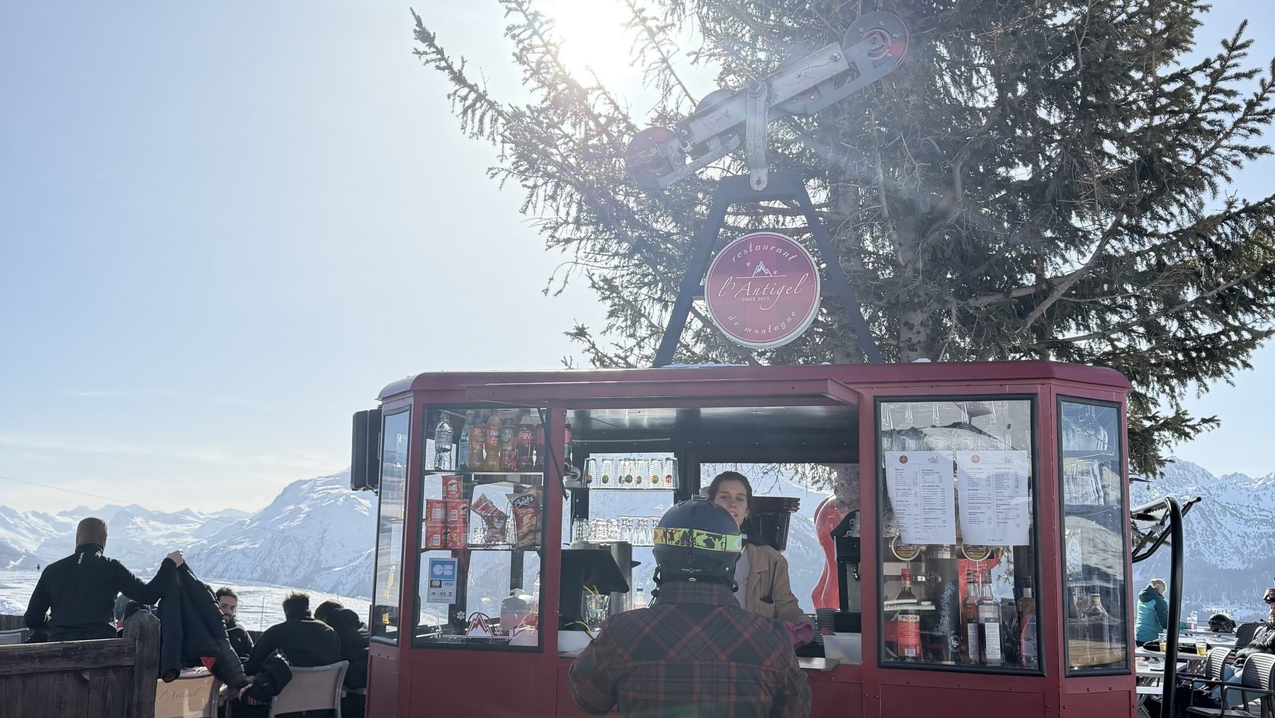 A red mountain café kiosk on a snowy terrace, with skiers nearby, an evergreen tree and snowy peaks beyond.