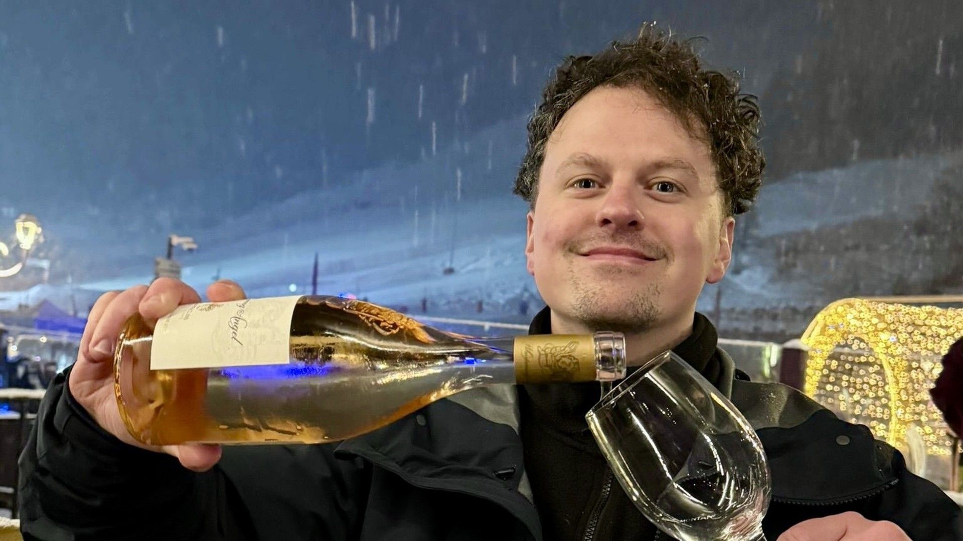 Smiling skier holds a large bottle of sparkling wine and a champagne flute in falling snow at night.