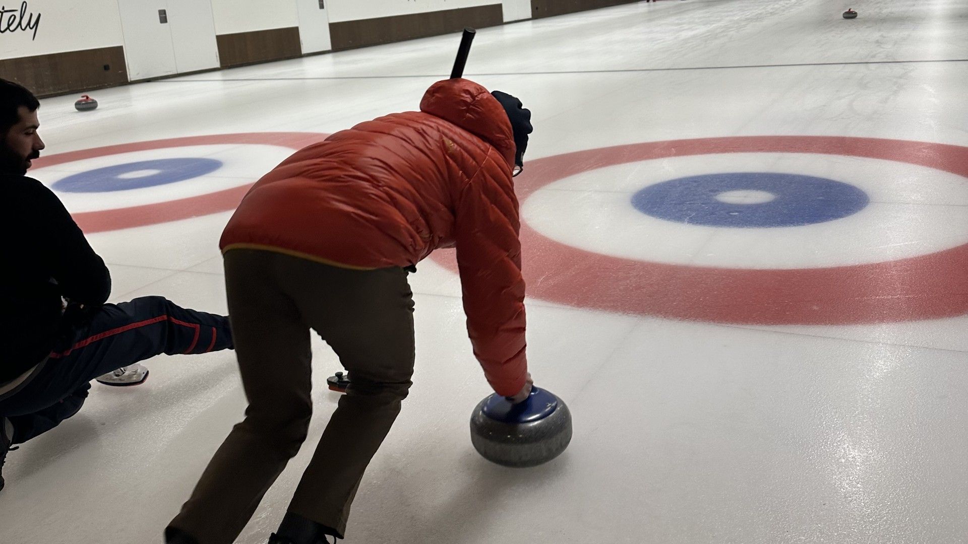 Skier bends low and slides a curling-style stone on an indoor ice rink