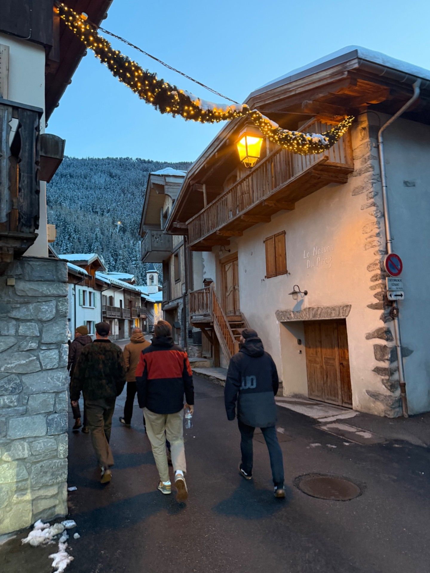 Friends walk through snowy Le Praz beneath festive lights, with Alpine chalets and a forested mountain behind them.