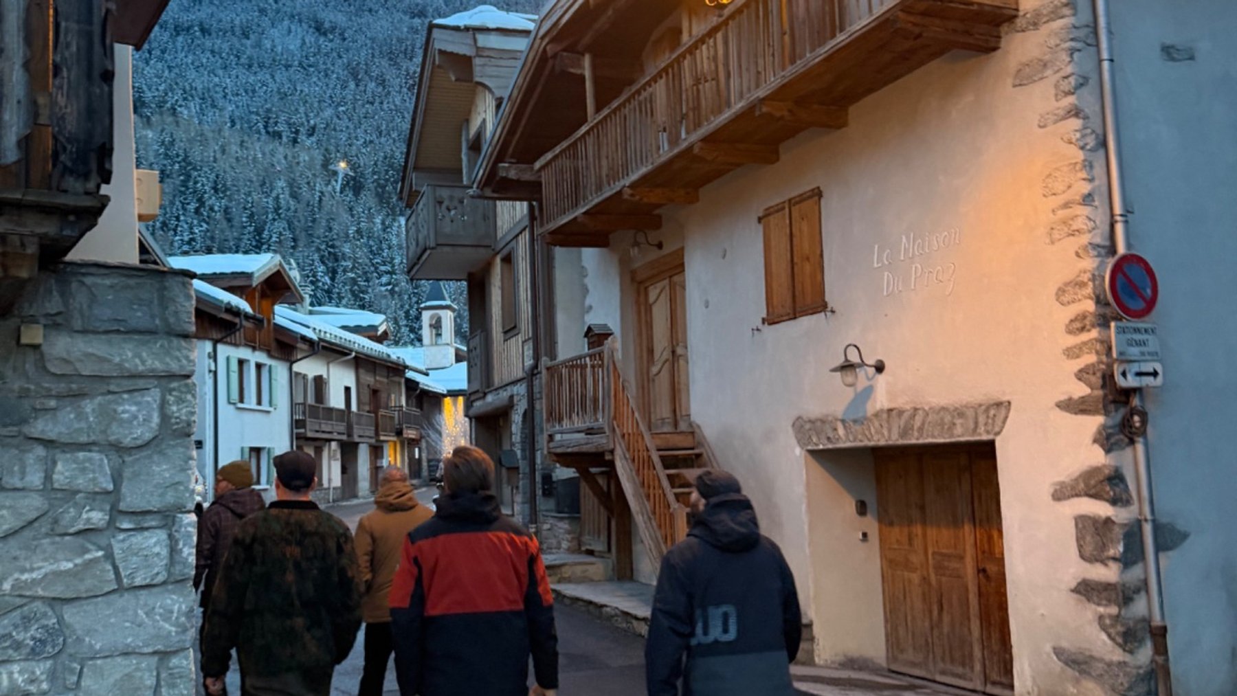 Friends walk through snowy Le Praz beneath festive lights, with Alpine chalets and a forested mountain behind them.