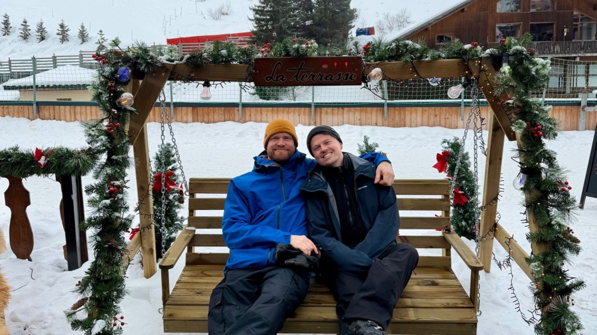 Two skiers sit on a decorated wooden swing outside a chalet in snowy Courchevel, with ski lifts behind them