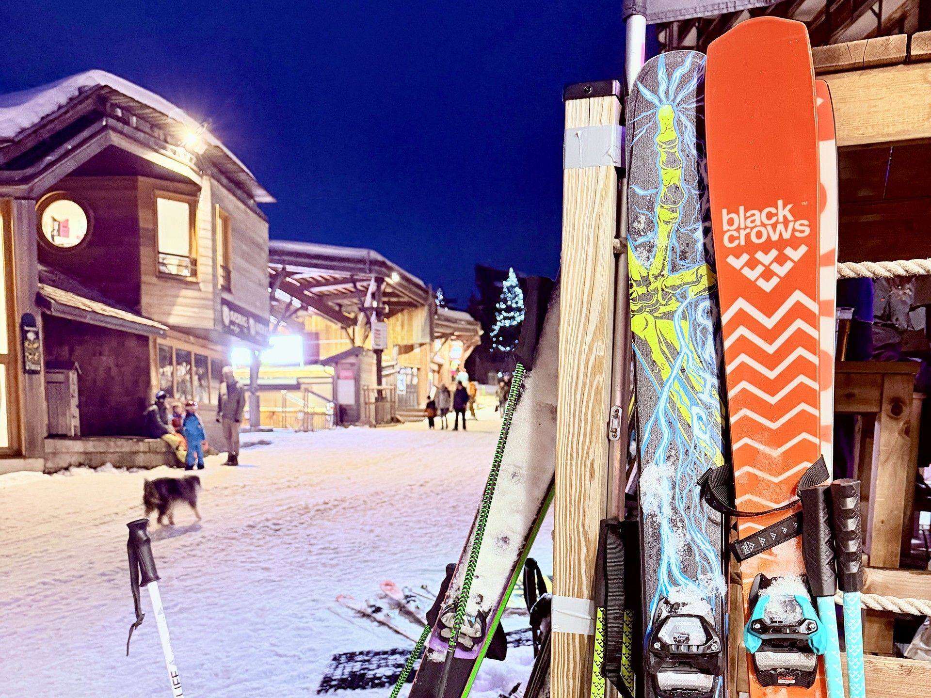 Ski lift poles and skis on a snowy resort path at night, with people skiing in the background.