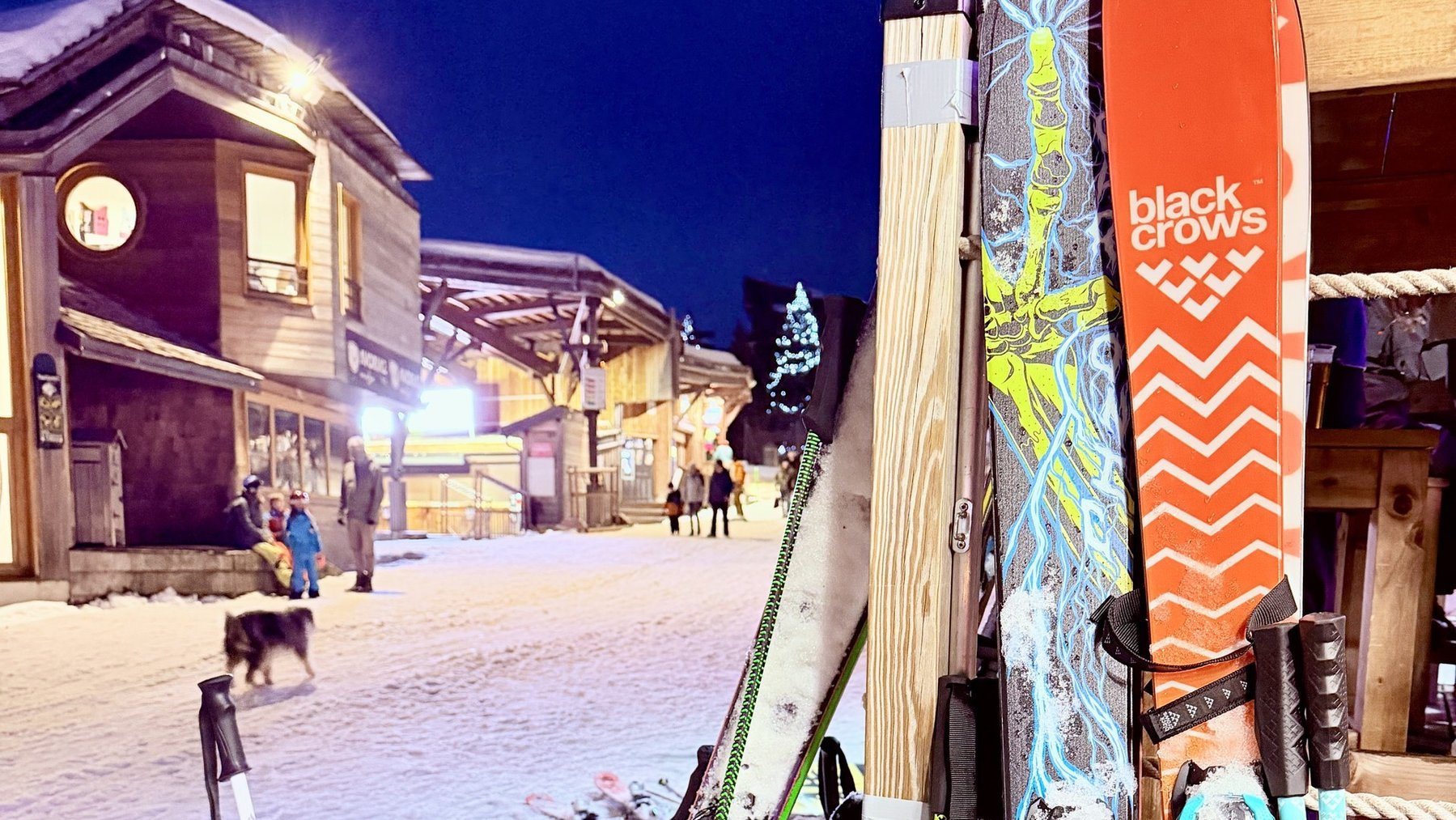 Ski lift poles and skis on a snowy resort path at night, with people skiing in the background.