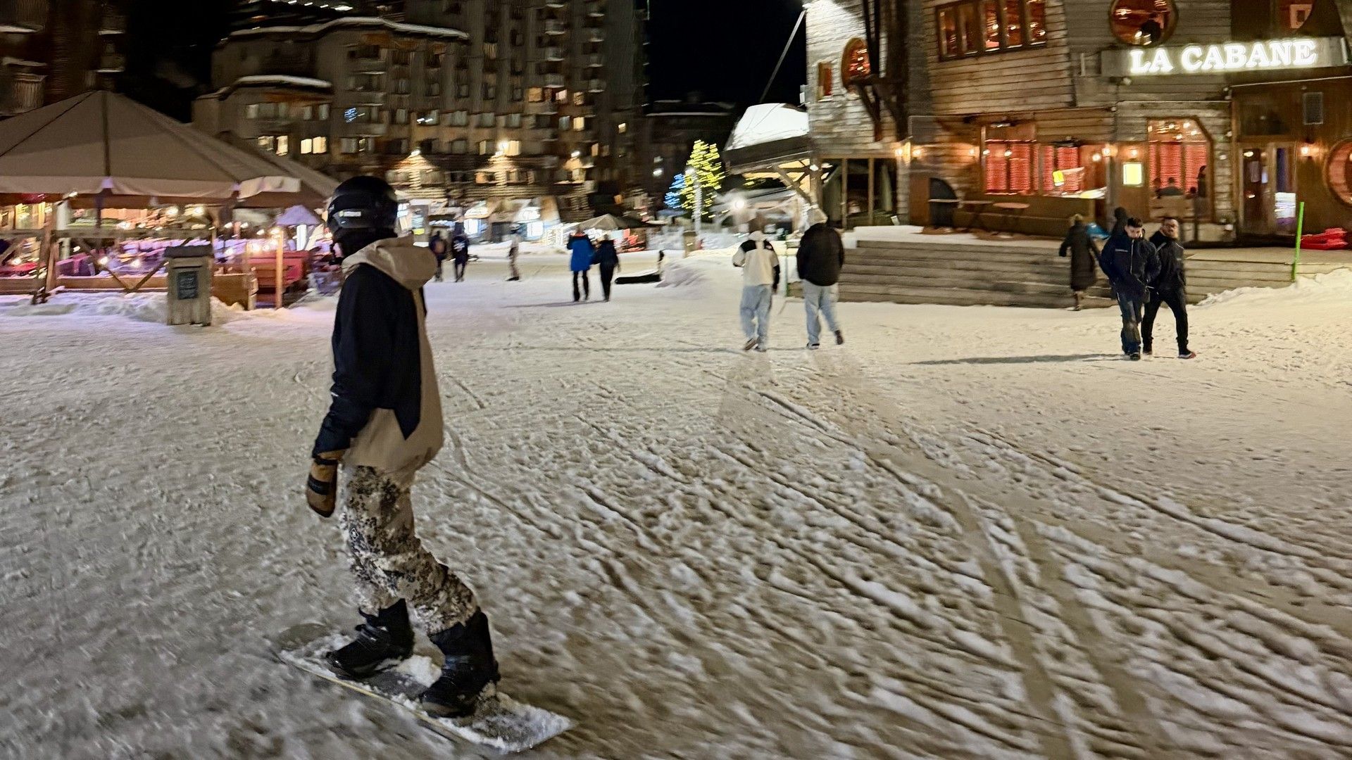 People walk through a snow-covered village square at night under bright winter lights