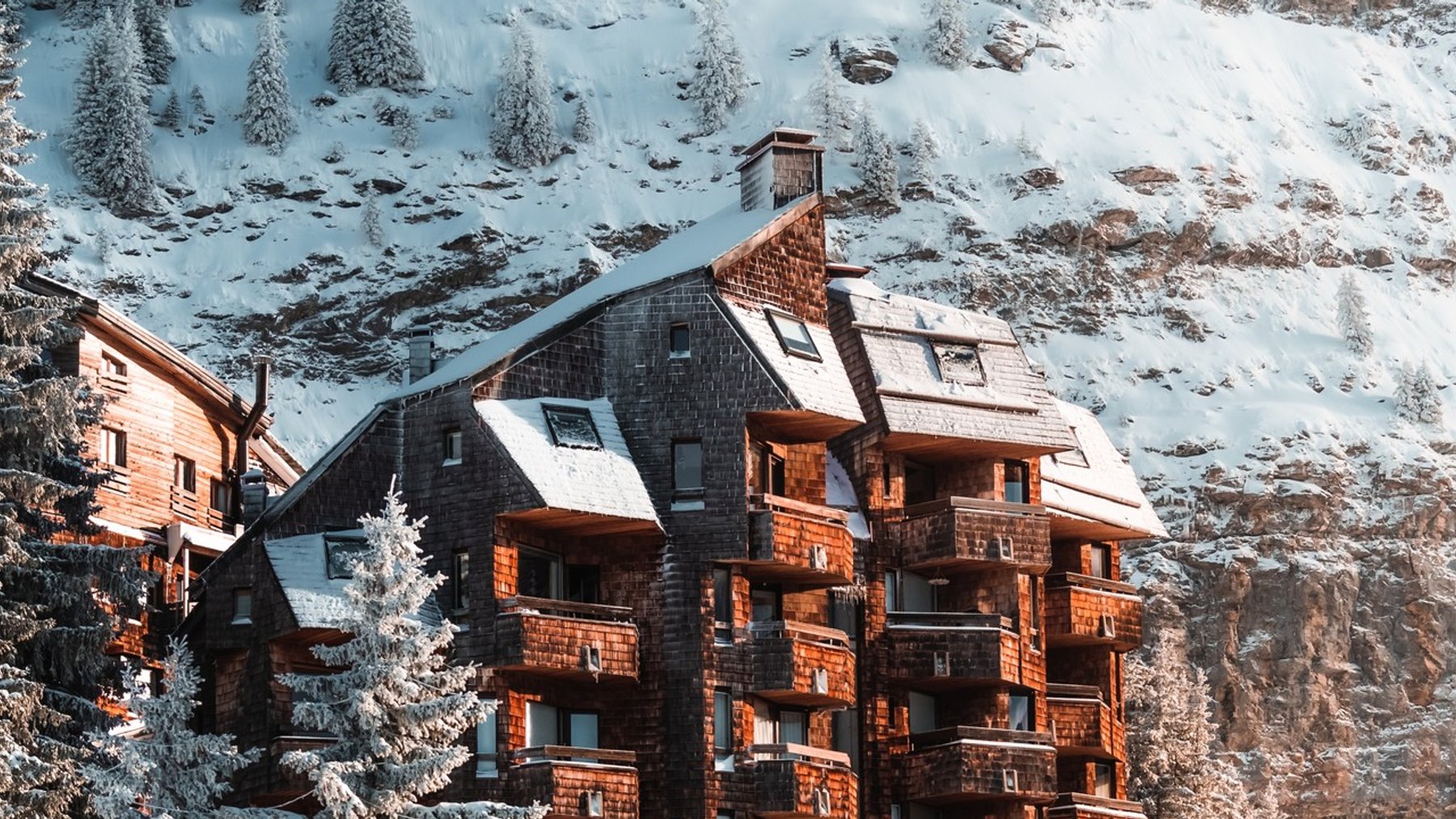 Snow-covered alpine resort buildings in a pine forest under a cloudy sky