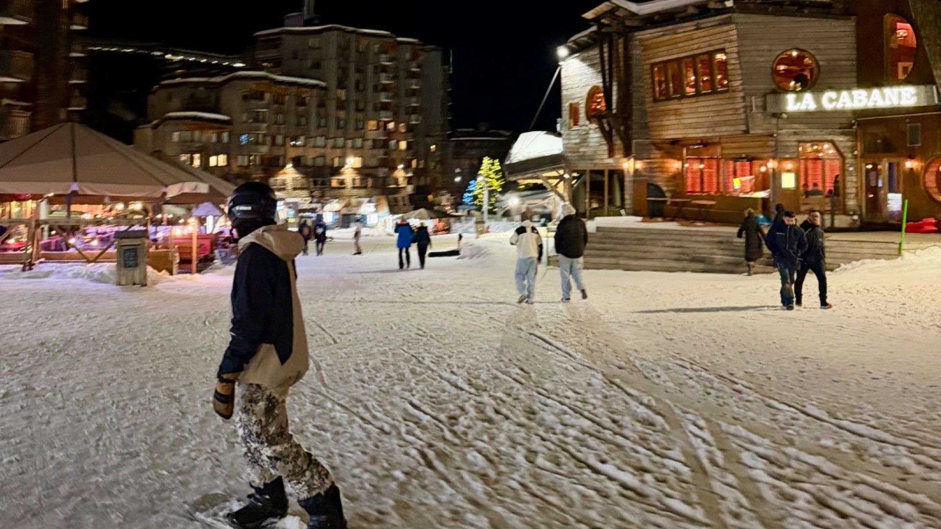 A skier crosses a snowy, floodlit square in Avoriaz, with resort buildings, pedestrians and a lit Christmas tree.
