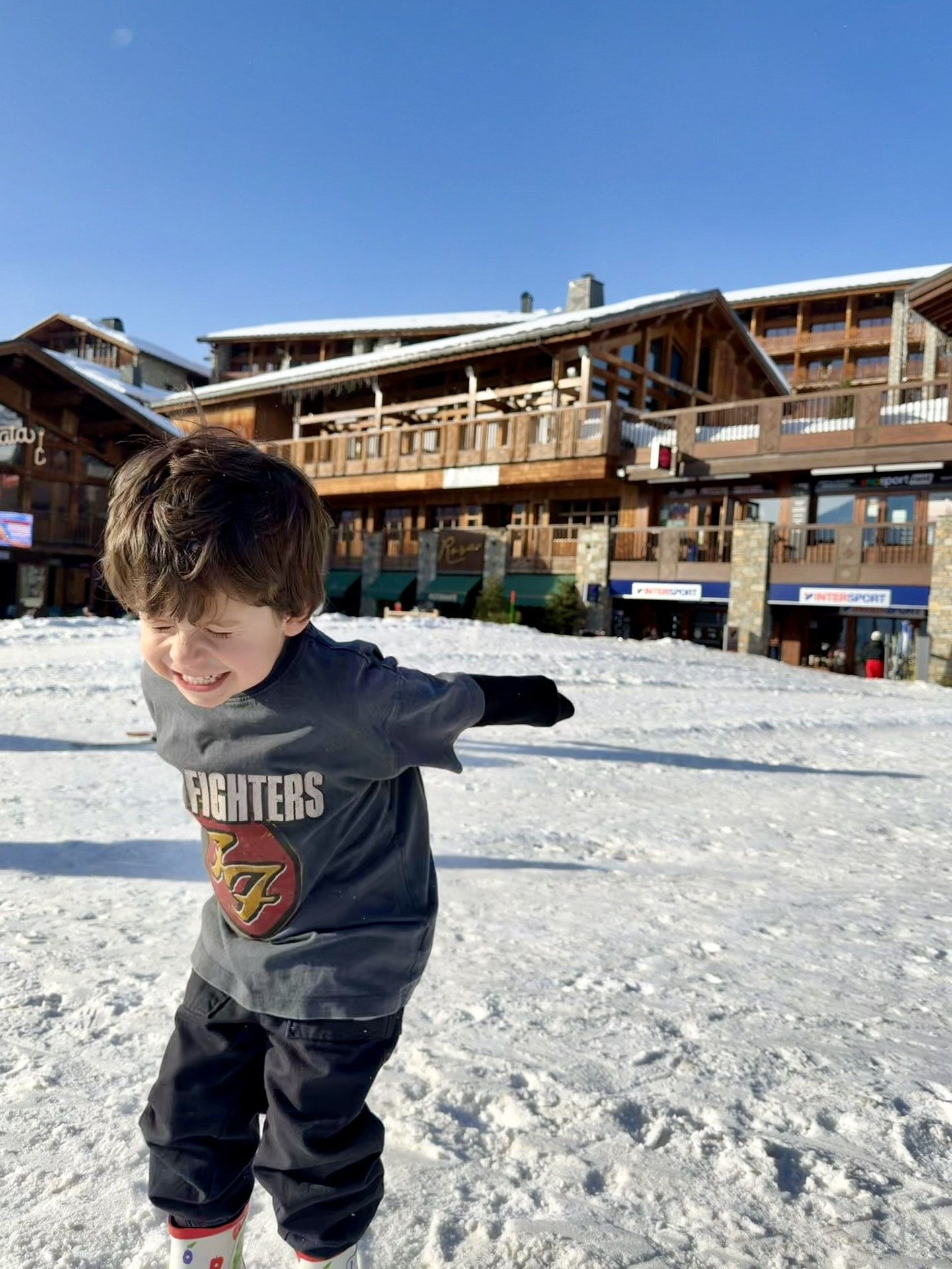 Child in ski gear points on a snowy slope outside a ski resort building