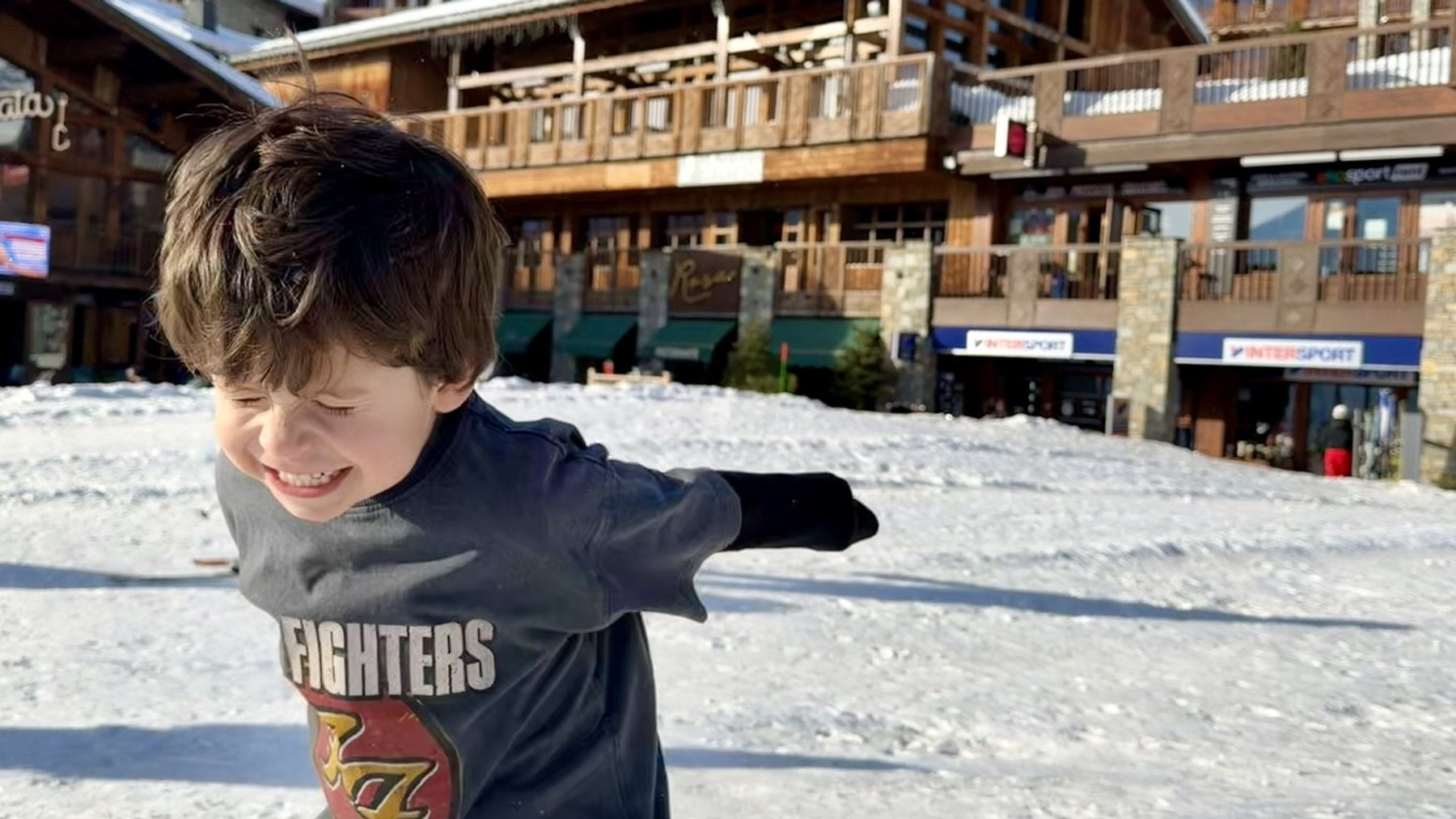 Child in ski gear points on a snowy slope outside a ski resort building