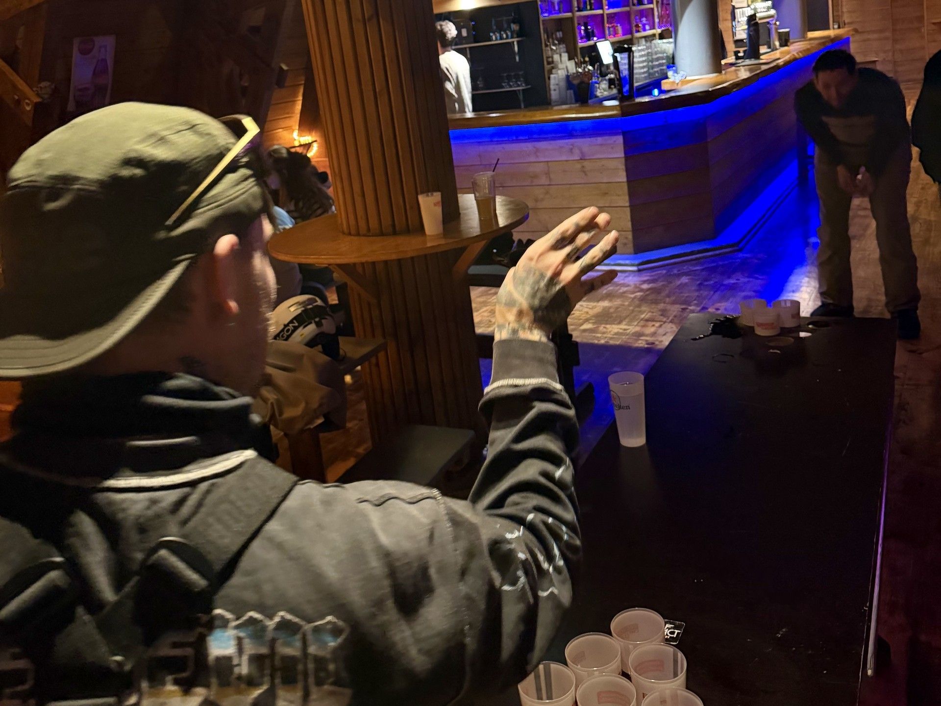 Person at a wooden bar counter reaches toward drinks in a dim ski lodge lounge with blue lighting