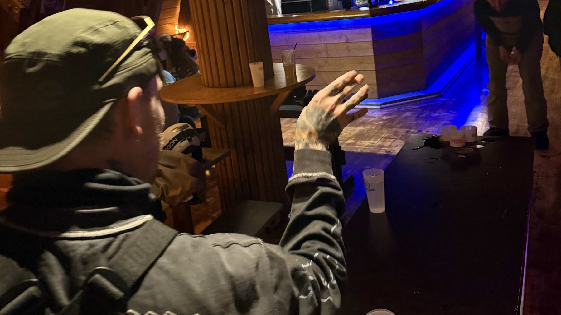 Person at a wooden bar counter reaches toward drinks in a dim ski lodge lounge with blue lighting