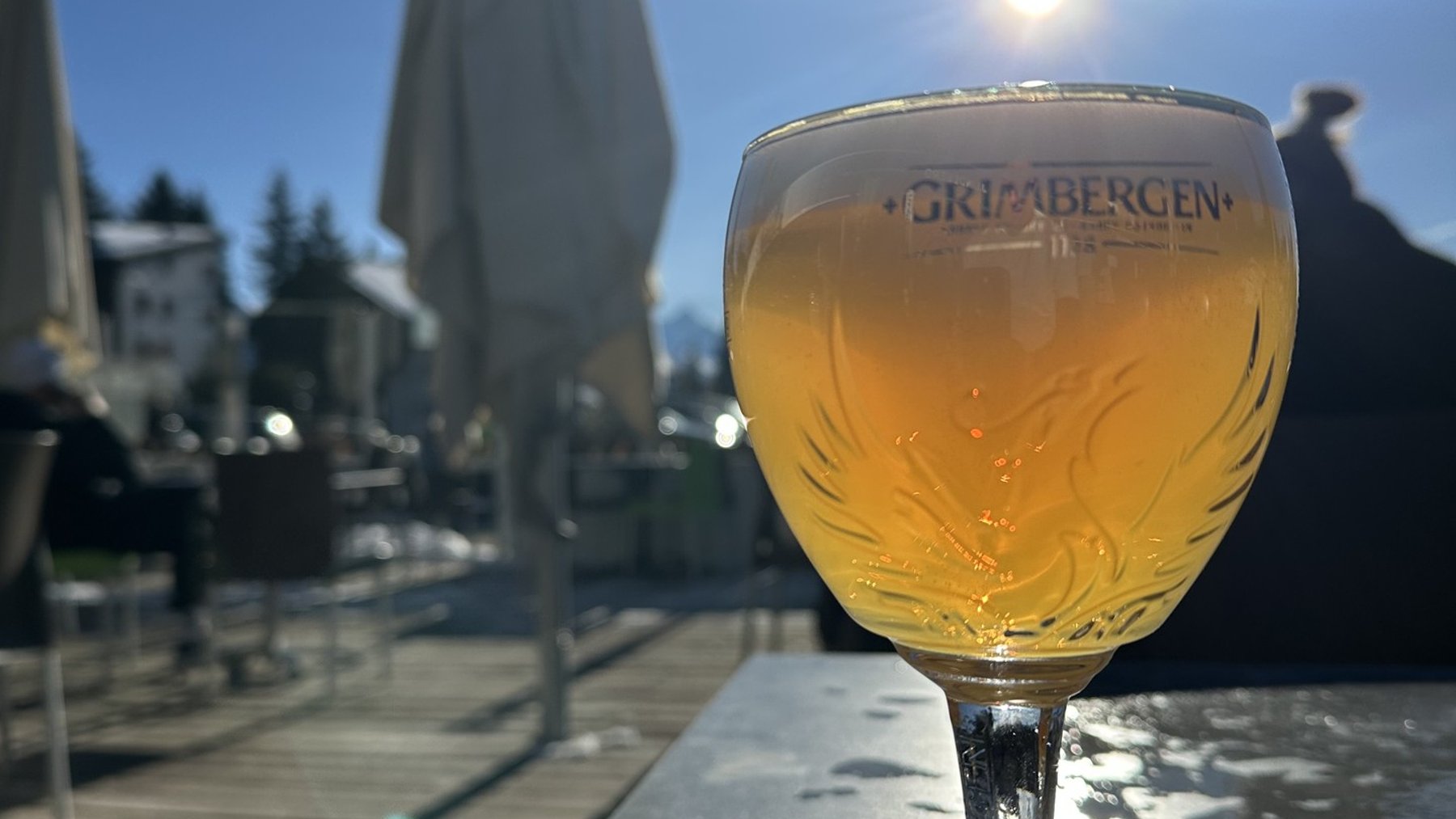 Golden beer in a glass on a lakeside terrace at sunset, with bright sun flares behind an umbrella