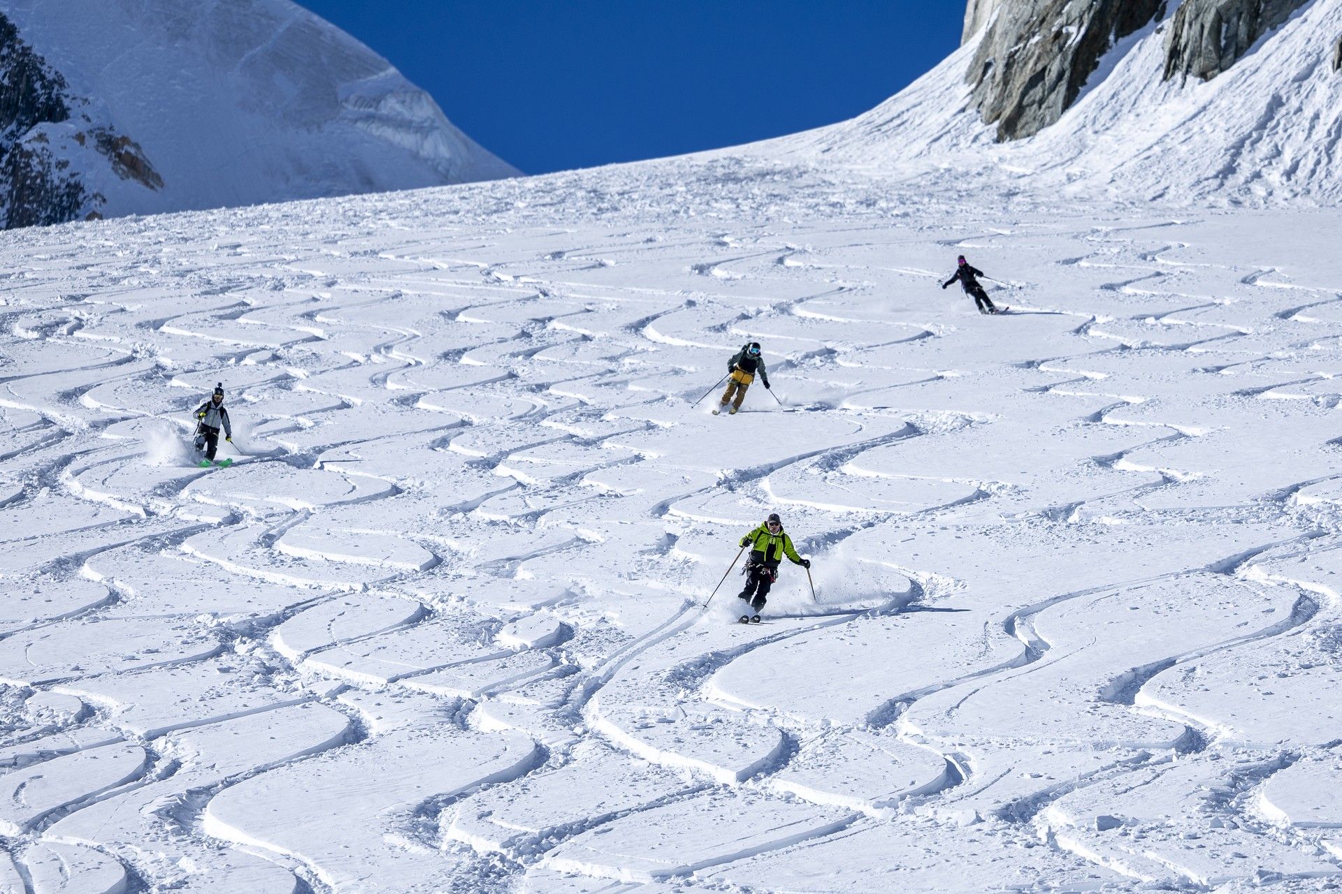 Skiers carve across a snowy alpine slope in bright conditions, leaving fresh tracks behind