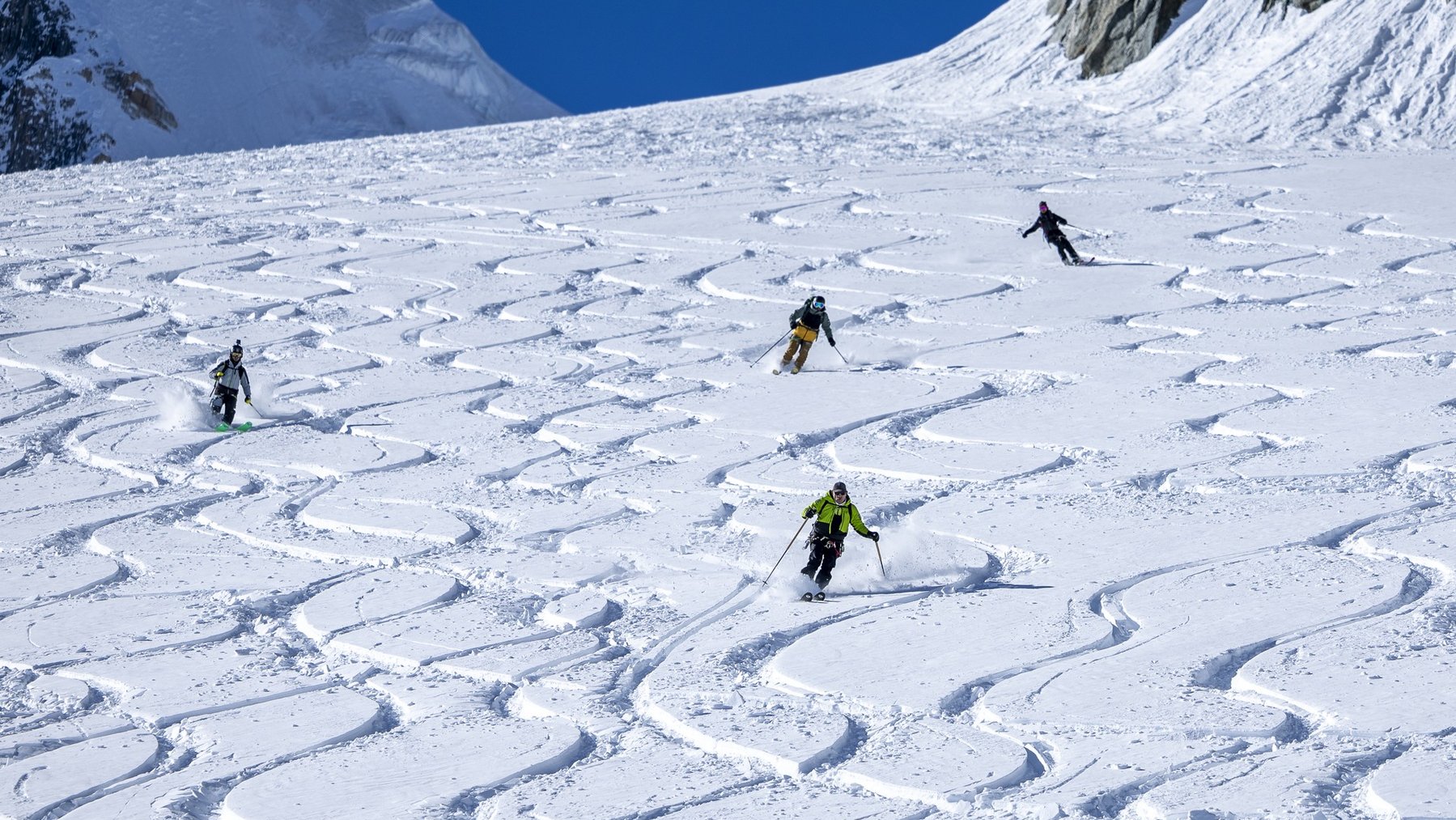 Skiers carve across a snowy alpine slope in bright conditions, leaving fresh tracks behind