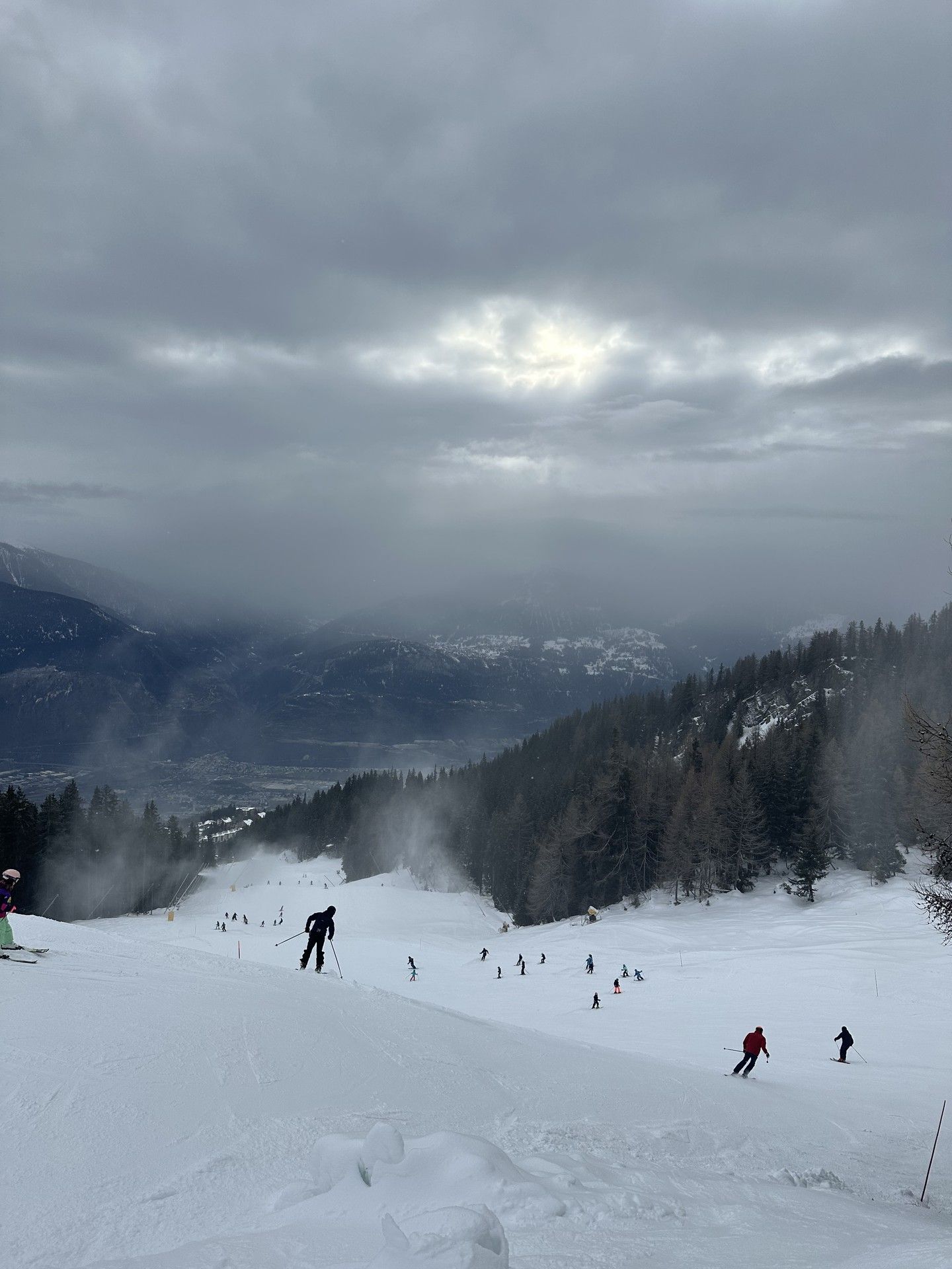 Skiers descend a snowy mountain slope under heavy clouds with a quiet valley view