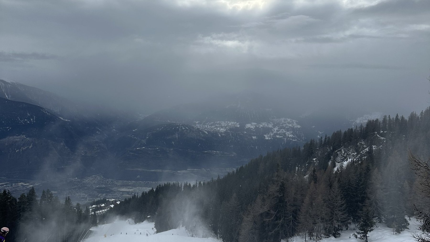 Skiers descend a snowy mountain slope under heavy clouds with a quiet valley view