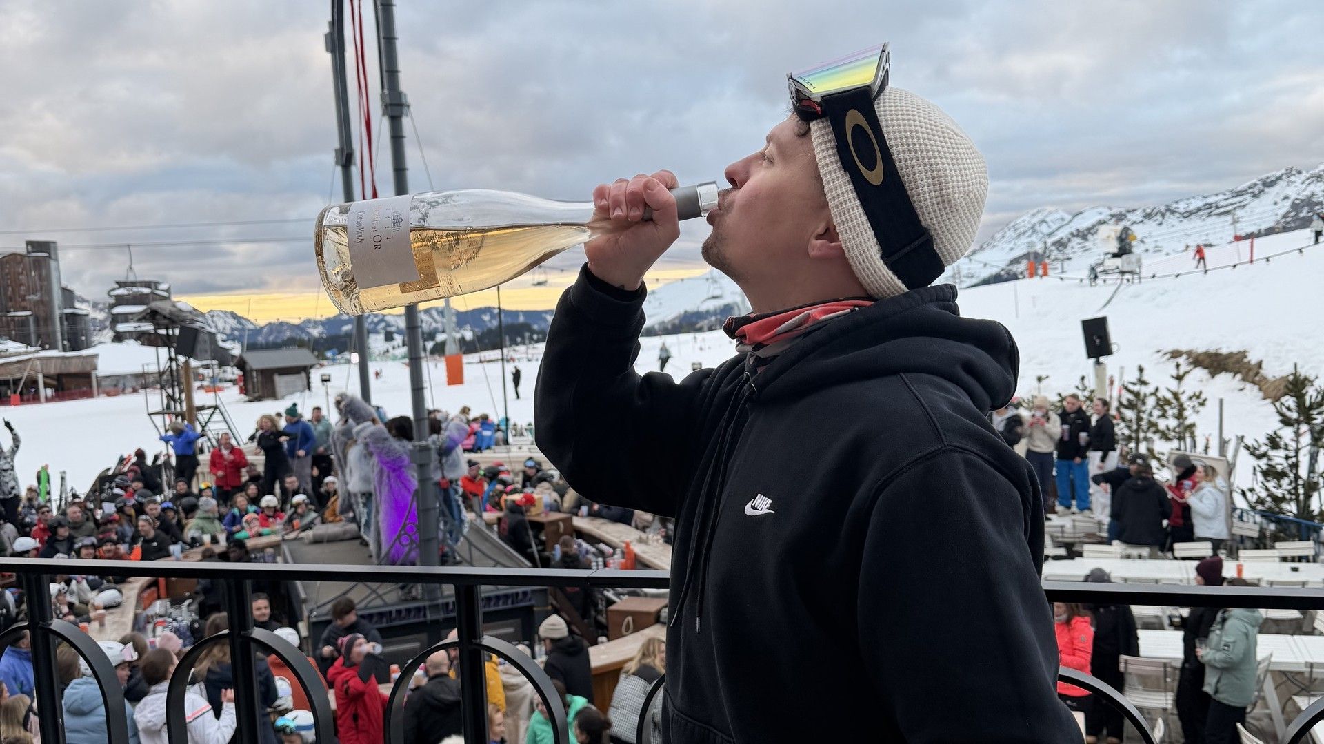 Ski spectator drinks from a paper cup at a resort viewpoint beside lift infrastructure on a cloudy day.