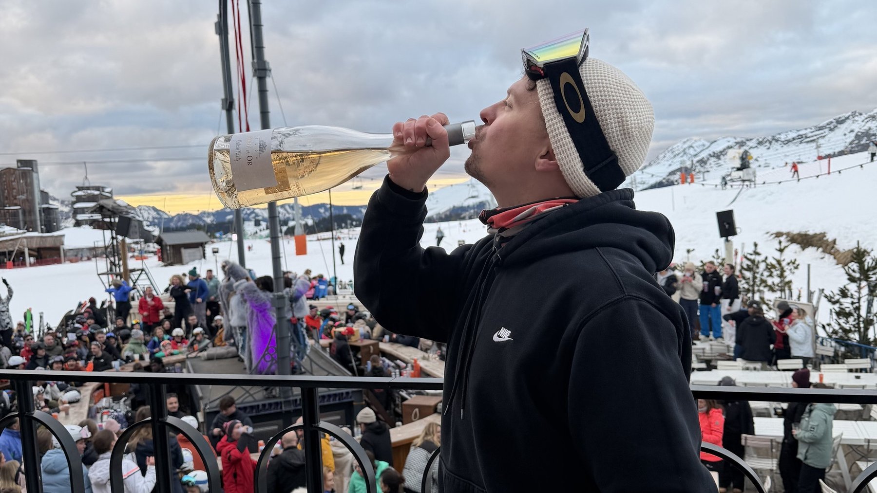 Ski spectator drinks from a paper cup at a resort viewpoint beside lift infrastructure on a cloudy day.