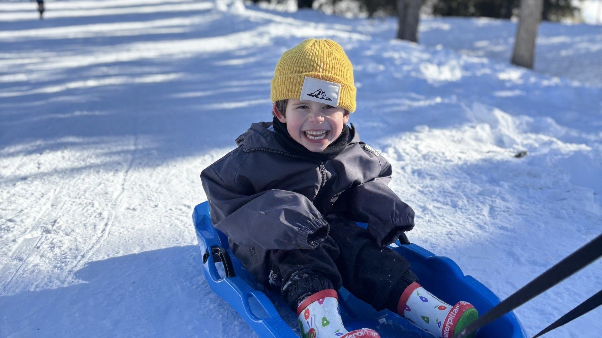 Child in a yellow hat rides a blue sled down a snowy slope in La Rosière