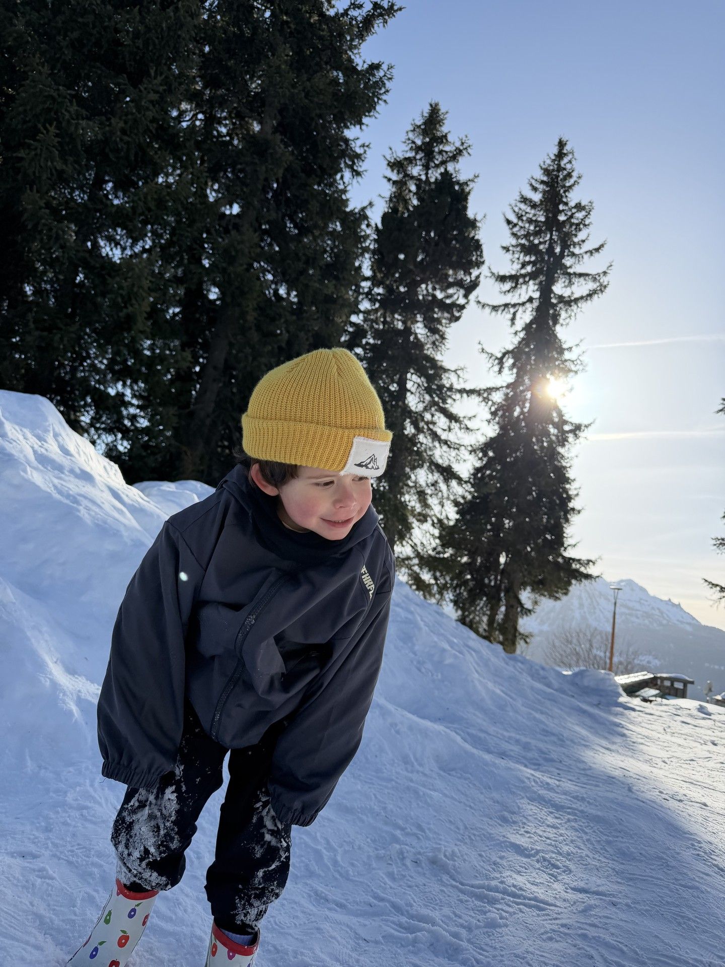 Snow-suited child in a mustard beanie and jacket standing on a sunny ski slope in alpine trees